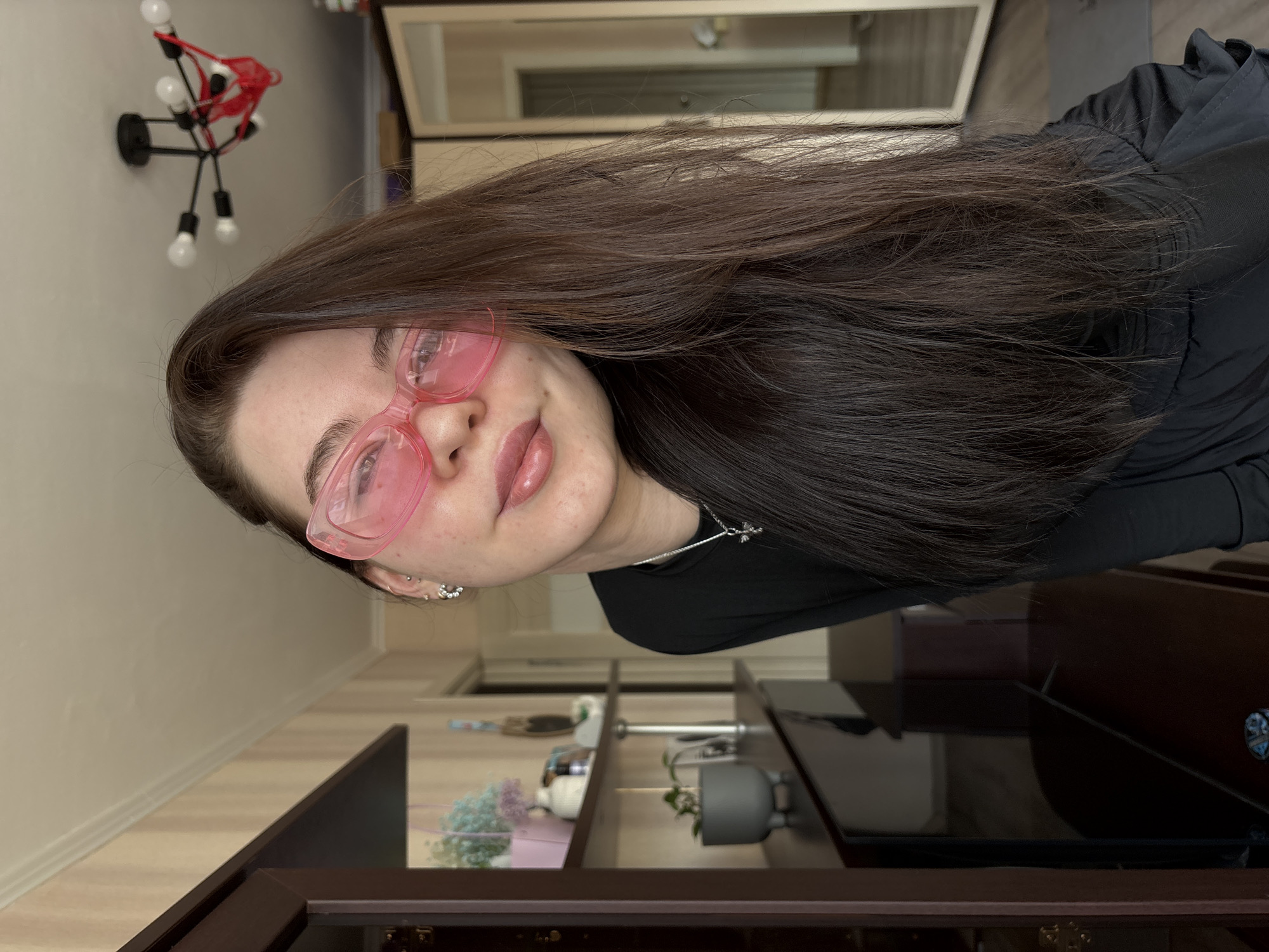 A young adult female is depicted in a close-up, chest-up view within an interior residential space in Perm, Russia. She has long, dark brown hair and is wearing oversized, translucent pink sunglasses, a dark long-sleeved top, a silver necklace with a cross pendant, and multiple silver earrings in her left ear. Her lips are glossy and slightly parted in a faint smile, revealing a dimple on her left cheek. The background on the left features a dark brown wooden cabinet displaying a pink box with artificial blue and purple flowers, a white ceramic object, and a small grey potted plant. The wall behind the cabinet has light-colored, vertically striped wallpaper. Overhead, a black, multi-bulb chandelier is suspended from the white ceiling, with a red thong draped over one of its arms. To the right, a large mirrored surface, likely part of a wardrobe, reflects an adjacent hallway, and a dark yoga mat is partially visible on a wooden floor.