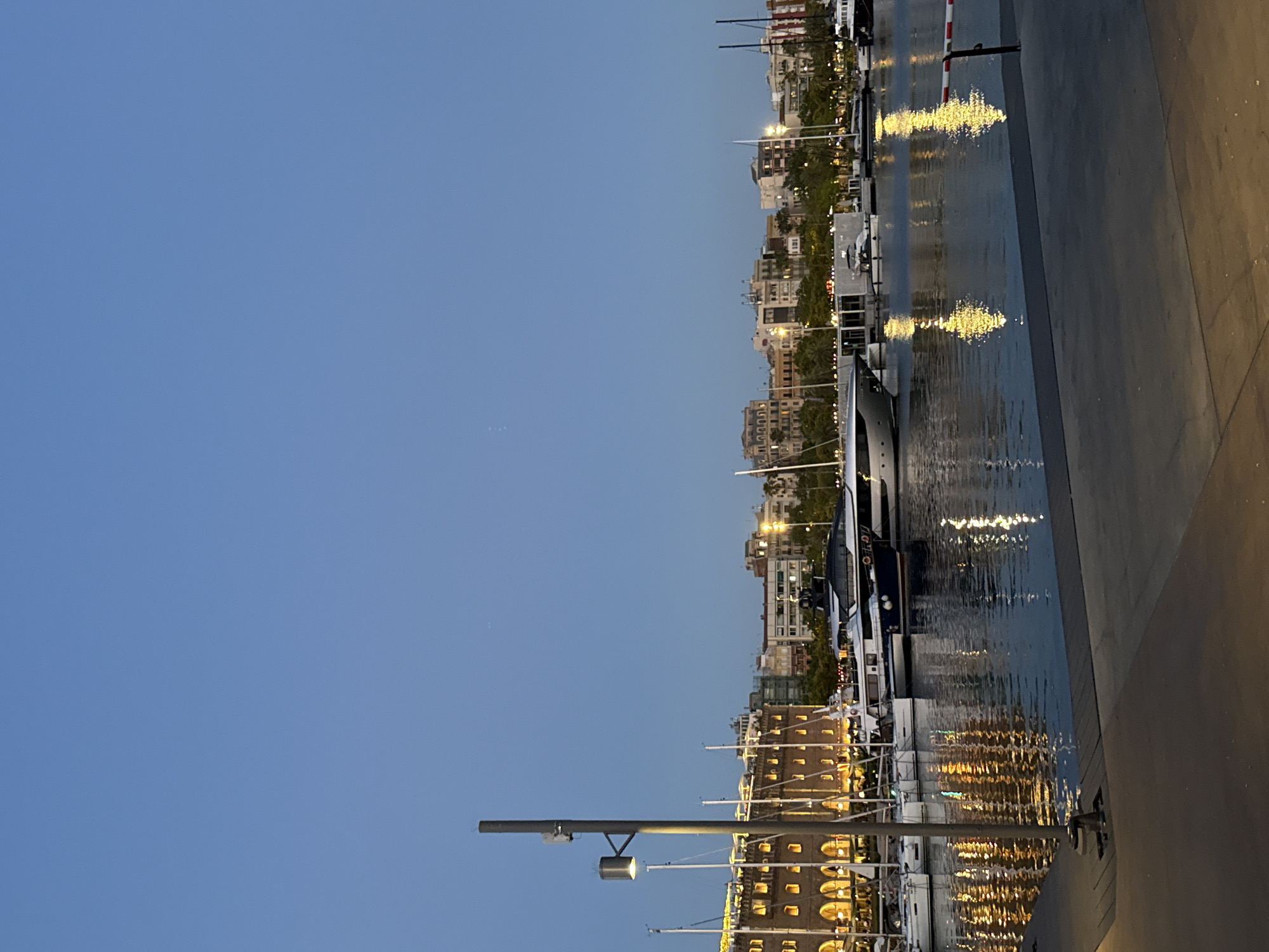 The image shows a harbor scene in Barcelona, Spain, at dusk. A promenade with a gray paved surface is in the foreground, with a bollard and a striped barrier post visible. The water of the harbor is calm, reflecting the lights of the city and the boats. Several yachts and sailboats are docked, with one large white yacht prominently featured. In the background, a row of buildings with illuminated windows lines the waterfront, characteristic of a city harbor. Palm trees are interspersed among the buildings, suggesting a Mediterranean climate. The sky is a clear, deep blue, indicating the transition from day to night. A tall, gray lamppost with a light fixture stands on the left side of the image. No people are visible in the scene.