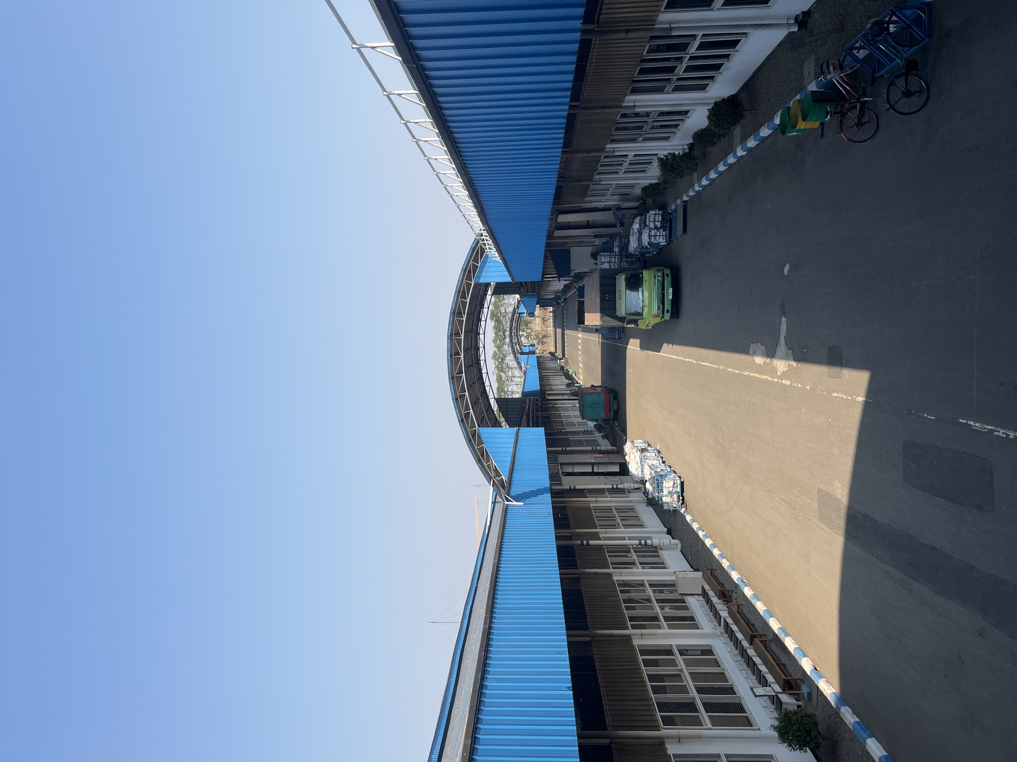 This is a daytime, eye-level, medium-shot photograph taken from an elevated perspective, looking down a covered walkway at what appears to be a loading or unloading area within an industrial complex. The location is identified as Pelang, Indonesia. The walkway is flanked by two long, low-rise buildings with blue corrugated metal roofs and brown metal siding. The buildings have numerous large windows with white frames. Along the left side of the walkway, there are wooden benches situated outside the building. On the right side, two green and yellow trash cans are positioned next to two bicycles, one red and one blue, leaning against a blue metal frame.
The walkway itself is paved with asphalt and has a white dashed line marking the center. Several stacks of white goods, possibly packaged materials, are organized on pallets and are visible on both sides of the walkway. A green truck is parked on the right side of the walkway, and another truck, also seemingly green, is further down on the left. The sky is a clear, pale blue. The scene suggests ongoing activity within a logistics or manufacturing facility.