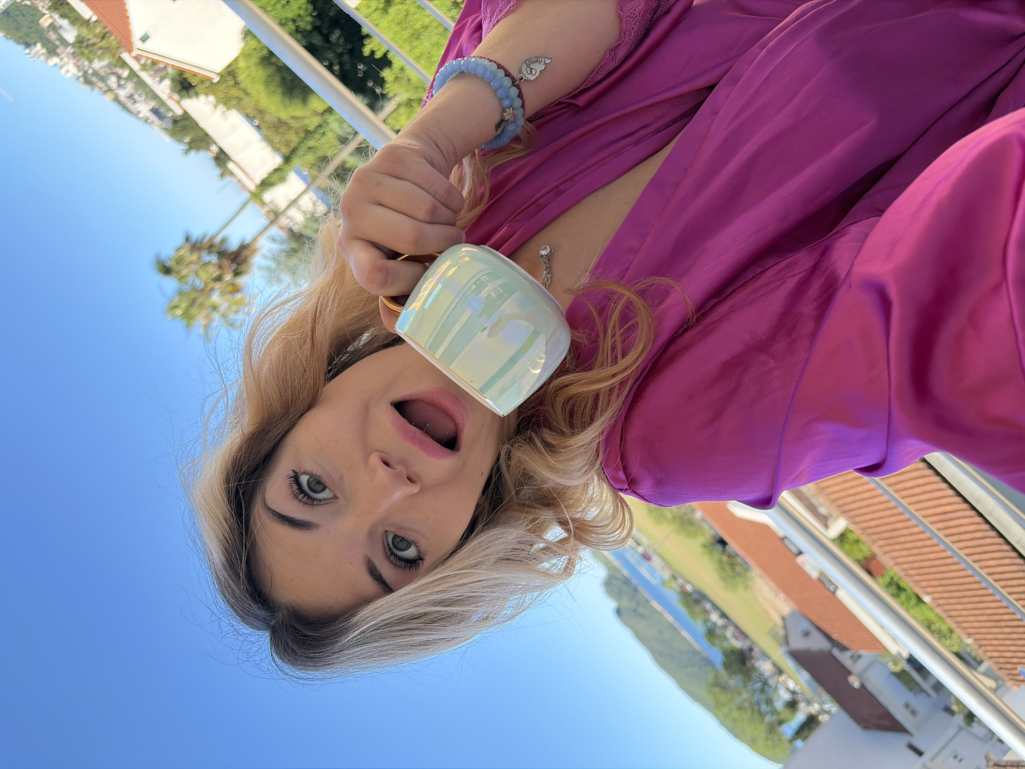 A woman in a bright pink robe holds a mug outdoors on a balcony in Budva, Montenegro. She is holding the mug to her mouth, and her mouth is open. She has blonde hair with dark roots and fair skin. She is wearing a bracelet and a necklace. The background shows a hillside with trees, rooftops of buildings, and the sea. The sky is blue. Balcony railings are visible at the side of the frame.