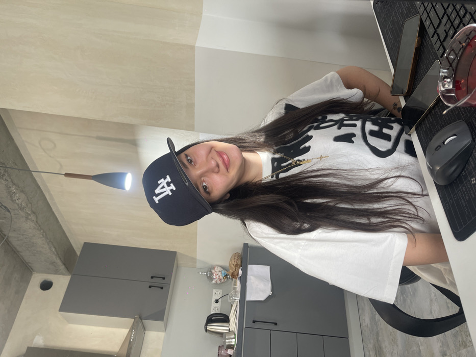 A female individual is seated at a desk in a modern interior setting, identified as a location in Yerevan, Armenia. She possesses long, dark hair and wears a black baseball cap featuring a white "LA" logo. Her attire includes a loose-fitting white t-shirt with dark graphic designs, and she accessorizes with a gold-colored chain holding a cross pendant and another small rectangular pendant. She is smiling subtly and looking directly forward.
The immediate foreground displays a light-colored desk surface. On the desk, from left to right, are a black wireless computer mouse positioned on a black mousepad, two smartphones laid face down (one with a dark case, one with a lighter-colored case), and a black computer keyboard. A clear glass mug containing a red liquid is situated to the right of the smartphones.
The background to the left reveals a contemporary kitchen area. It features dark grey upper and lower cabinets with black handles, a silver kettle, and a silver range hood. A white power outlet is visible on the wall above the counter. A tall glass jar filled with colorful candies, a draped white cloth, and a brown paper bag are also present on the counter. The walls are painted in a two-tone scheme of light beige/off-white, topped by an exposed concrete ceiling from which a dark-colored pendant lamp with a wooden accent hangs, emitting a downward light.