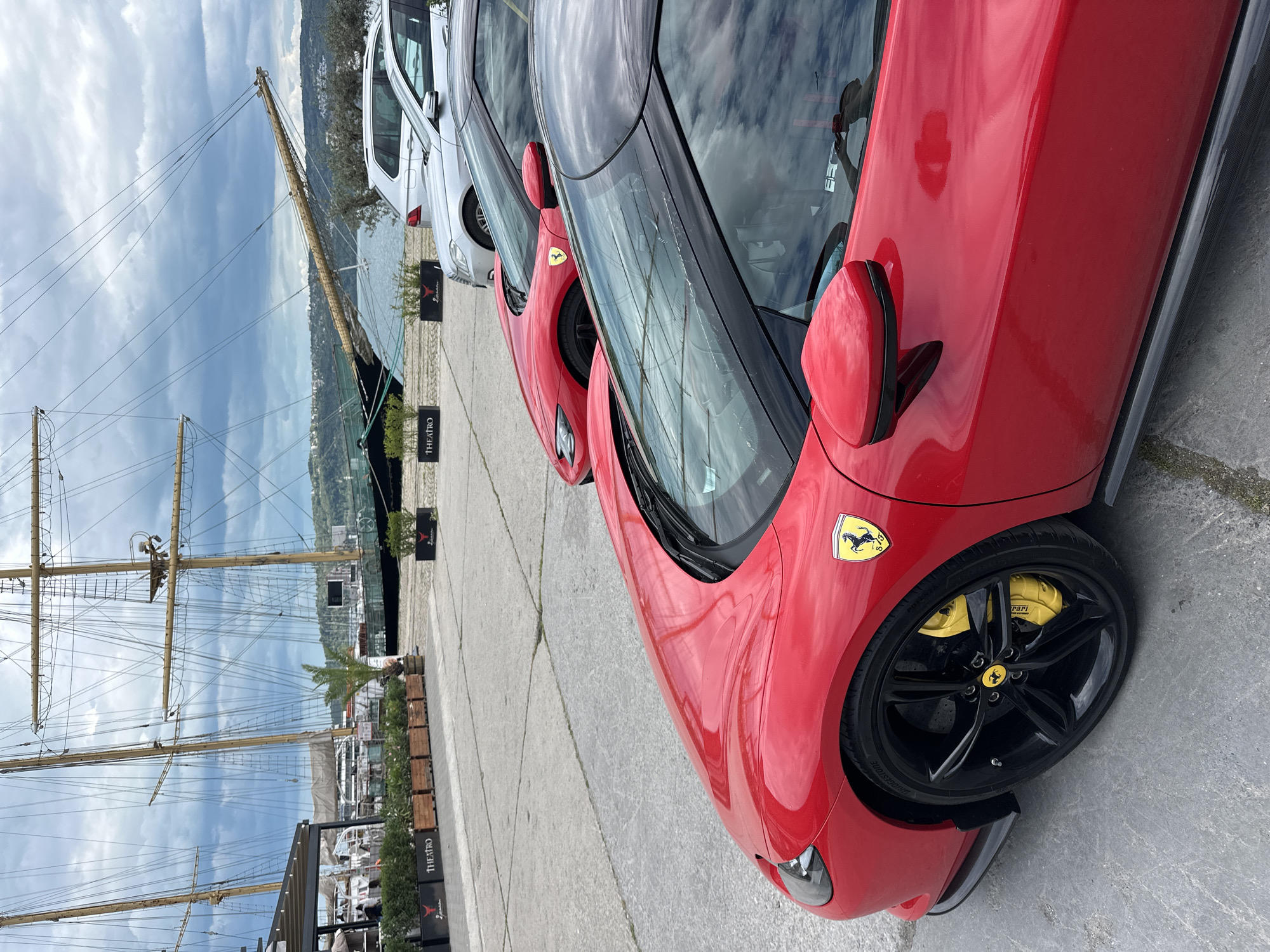 The image shows a row of parked vehicles, predominantly red Ferrari sports cars, along a paved waterfront area. To the left of the cars, the masts and rigging of a large sailing ship are visible, indicating a maritime setting. In the background, a body of water with a hilly shoreline can be seen. The sky is overcast, suggesting a cloudy day. The presence of the sailing ship and the overall setting are consistent with the city of Varna, Bulgaria, which is known for its historical ties to maritime activities and its location on the Black Sea coast. Additional clues in the background suggest the presence of shops or businesses that are near the waterfront. The "TEATRO" sign, which is partially visible, hints at the location near a theater or performing arts venue.