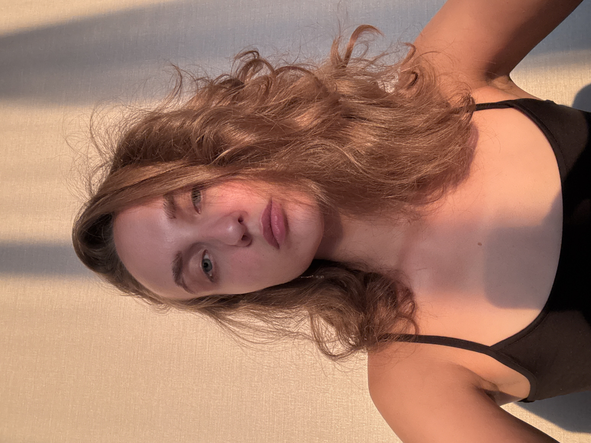 A young woman with wavy, light brown hair is the subject of this portrait. She is wearing a black tank top. Her head is tilted, and she is looking directly at the camera. Her eyes are blue-green, and she has light pink lips. The background is a textured wall, with a diagonal shadow cast across it from the left side of the frame, suggesting sunlight. The lighting and shadow indicate it is likely daytime, possibly late afternoon or early morning. The image is oriented vertically, with the woman's head towards the left side of the frame. The setting appears to be indoors, possibly in a room in Podshivalovo, Russia. There is no visible text in the image.