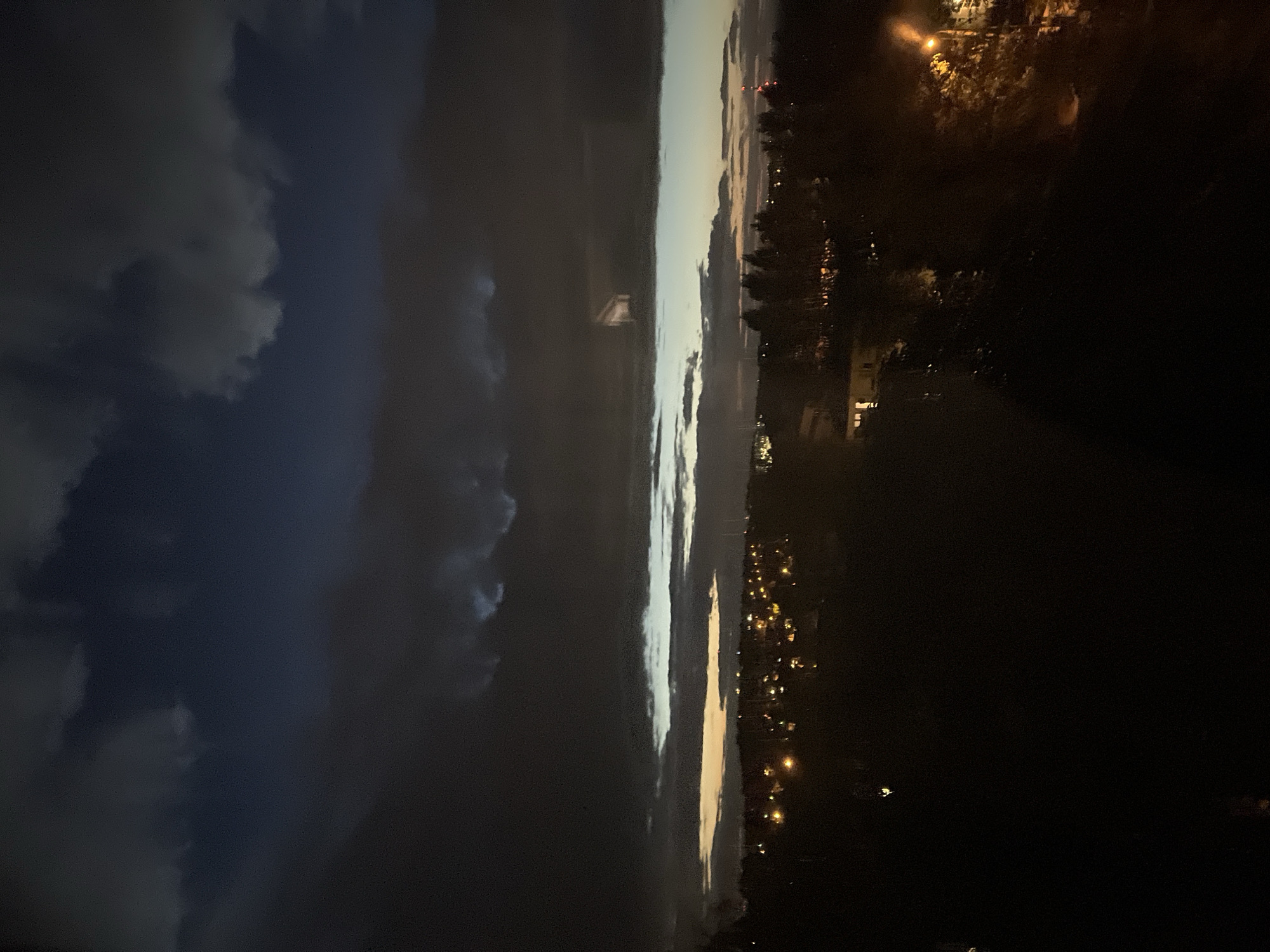 An aerial view captures a twilight scene over a dark, tree-lined landscape dotted with illuminated structures in the distance. The sky above is a gradient of deep blues and grays, with dramatic, heavy clouds obscuring much of the light. A faint, rectangular, light-colored object is visible within one of the clouds, appearing to be suspended in the atmosphere. The lights of a settlement twinkle below, suggesting a human presence. The foreground is dominated by deep shadows, rendering details of the terrain indistinct. A solitary streetlamp illuminates a portion of the right side of the image with its warm glow.