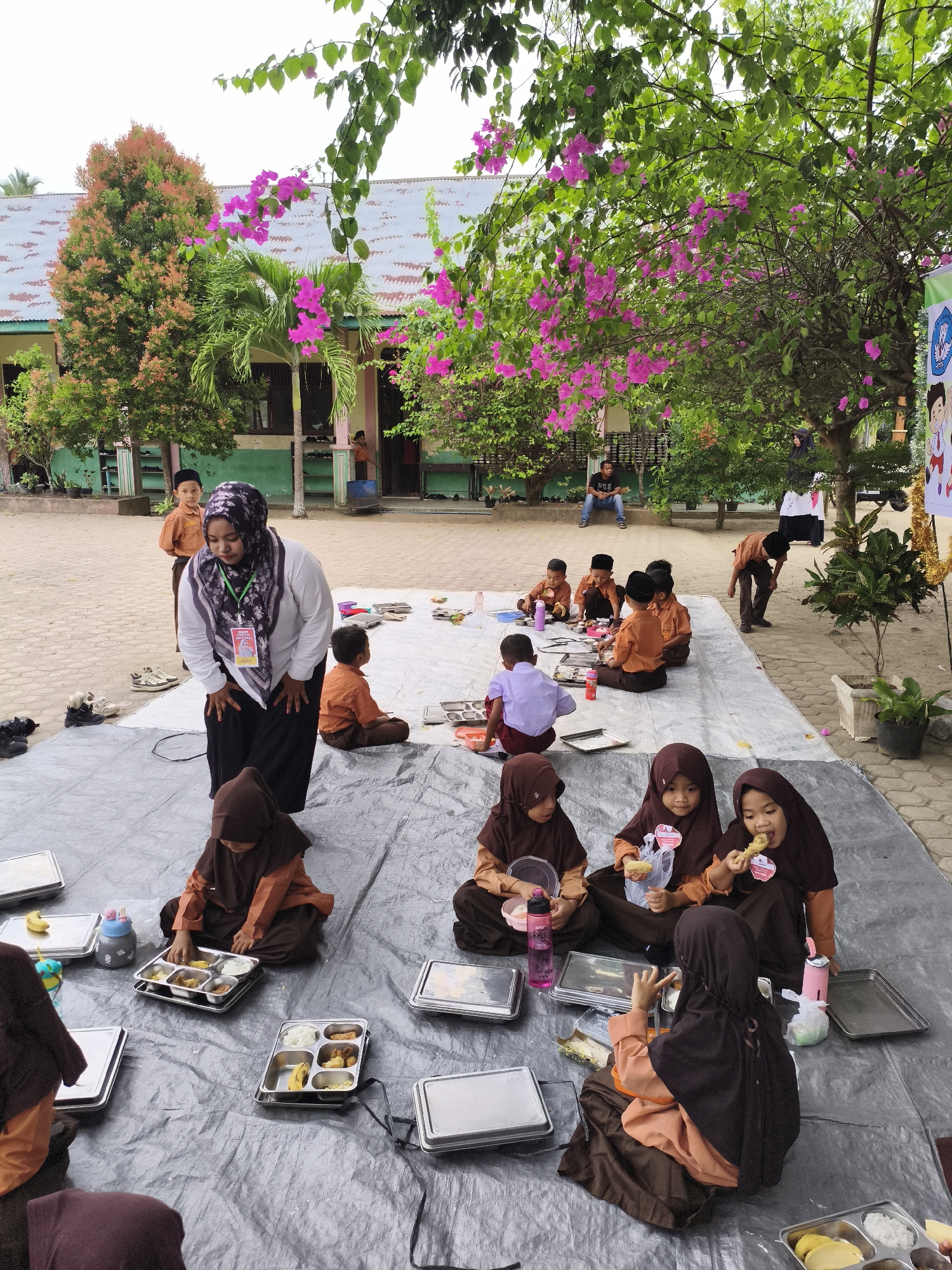 The image depicts an outdoor scene in what appears to be a schoolyard in Baratpayaitek, Indonesia. A group of children, mostly dressed in school uniforms consisting of brown tunics and headscarves, are seated on a large gray tarp laid out on the ground. They are engaged in eating from metal lunch trays that contain rice, fruit, and other food items. Some children are actively eating, while others are in the process of serving themselves or interacting with their food.
A woman, likely a teacher, is standing and bending over near the children, possibly supervising or assisting them. She is wearing a white shirt with a black headscarf adorned with a colorful pattern and a badge. In the background, more children are visible, some standing and others seated. Buildings with light-colored walls and metal roofs are present, along with trees and flowering plants, including vibrant pink bougainvillea. The ground surface appears to be paved. A man is seated on a bench in the background, observing the activity. The overall atmosphere suggests a school event or mealtime.