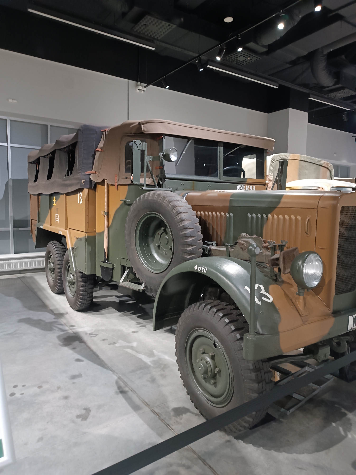 The image shows a vintage military truck, likely from the mid-20th century, on display indoors. The truck is painted in a camouflage pattern of brown and green. It has a canvas-covered rear cargo area, a spare tire mounted on the side, and a prominent front grille. The vehicle is situated on a concrete floor, and the background reveals an exhibition hall with industrial-style lighting and ceiling structures. To the left, a large window with vertical blinds is visible. The truck is positioned with its left side facing the camera. The scene appears to be in the Museum of Military Technology "Battle Glory of the Urals" in Verkhnyaya Pyshma, Russia, given the context of such exhibits. There are no people visible in the image, and the truck is stationary.