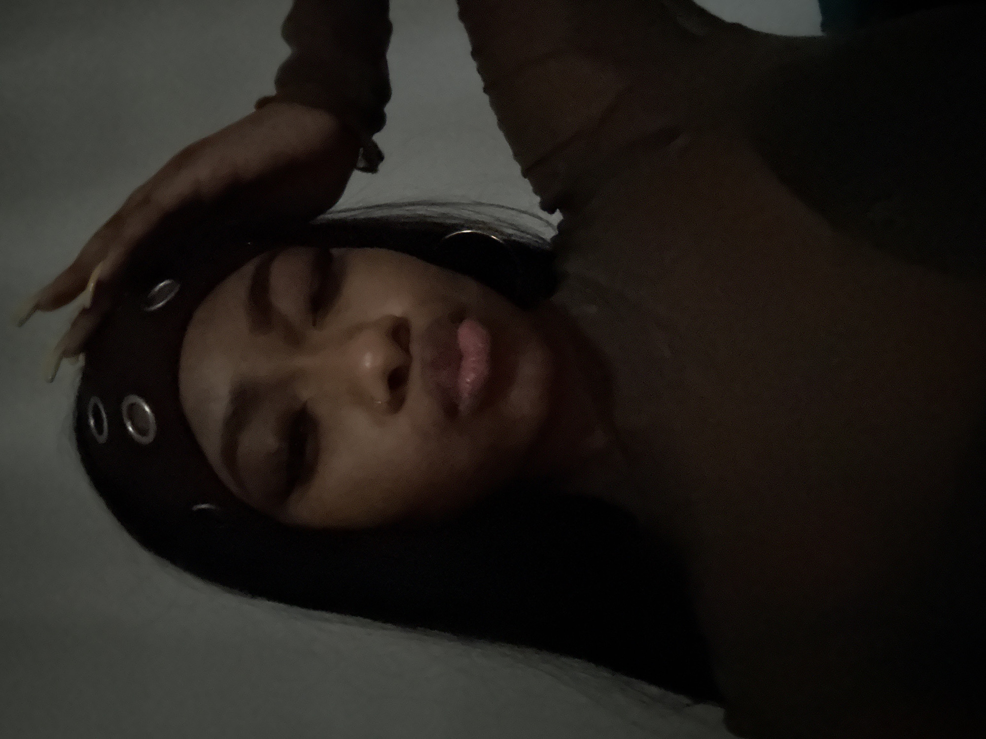 The image shows a person with dark hair and dark skin lying down. They are wearing a headband with metal grommets. The person's eyes are closed, and their lips are slightly parted. Their right hand is raised near their head. The person appears to be wearing a brown shirt. The background is dimly lit and blurry, with a light-colored surface visible. The image is taken from an overhead perspective. There is no evidence to suggest that this image was taken in the city of Troitsk, Russia.