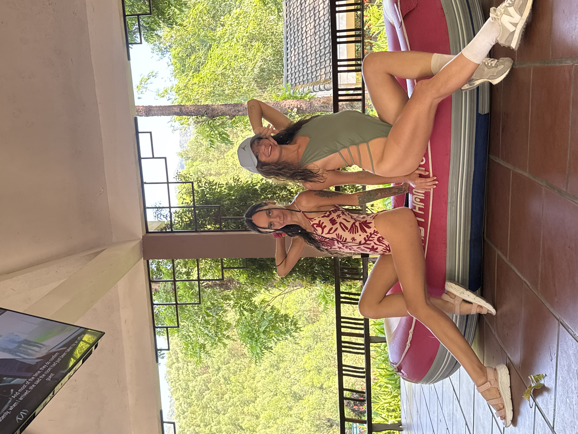 Two women are seated on a red inflatable boat on a tiled patio or porch. The woman closer to the camera is wearing a red and beige patterned one-piece swimsuit. The other woman is wearing an olive-green one-piece swimsuit and a gray baseball cap. Both are posing with their hands near their heads. Behind them is lush greenery. The location is the city of Đà Nẵng, Vietnam.