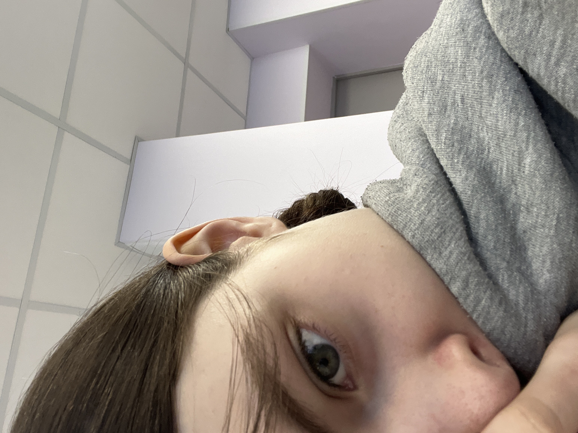 A close-up, angled view of a person's face and upper body. The person has fair skin, dark brown hair, and a green eye is visible. The person is partially obscured by a gray, textured fabric garment. The background reveals a white tiled wall and a white architectural element, potentially a ceiling or part of a structure, with what appears to be a section of a doorway or window opening. There is no information in the image to suggest that this photograph was taken in Saint Petersburg, Russia.