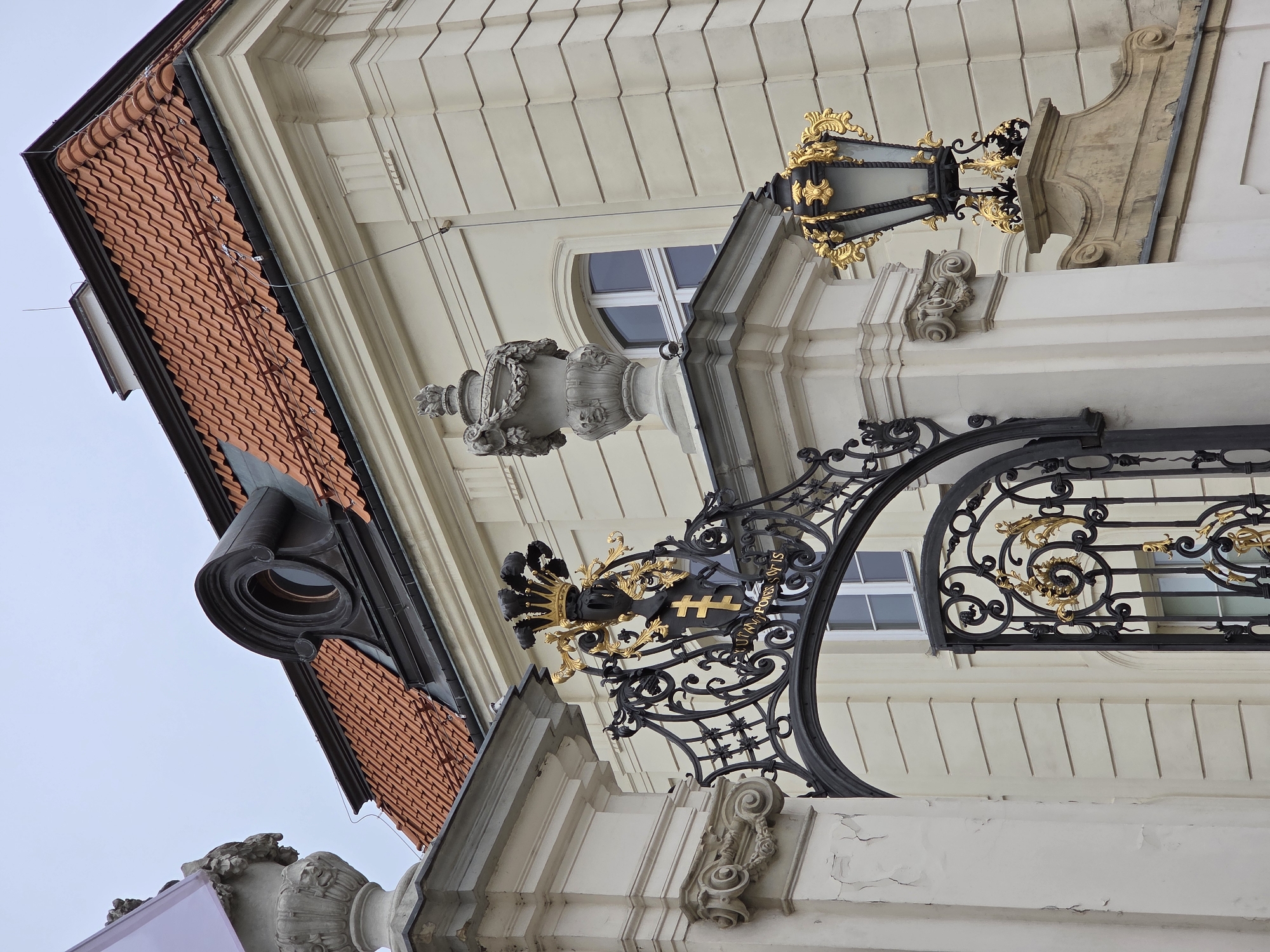 The image captures a detailed view of architectural elements in Warsaw, Poland. Visible features include the tiled roof and ornate facade of a building. A decorative black and gold wrought-iron gate stands prominently, adorned with intricate designs and a heraldic shield bearing a cross. Above the gate, an antique lantern is mounted on the building's facade, its gold embellishments contrasting with the pale building exterior. The architectural style suggests a historical or classical building, with decorative molding and stone embellishments enhancing the aesthetic. The angle of the shot looks upward, emphasizing the building's height and decorative features.