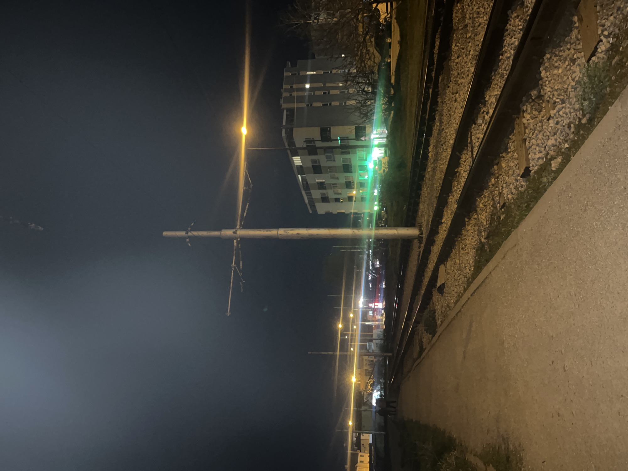 At night in Sofia, Bulgaria, the image captures a view along a railway track. To the right, there are railway tracks and gravel. A concrete walkway runs parallel to the tracks. Overhead, wires are supported by metal poles. In the distance, there is a tall building and a line of streetlights. The night sky is dark. No people are visible.