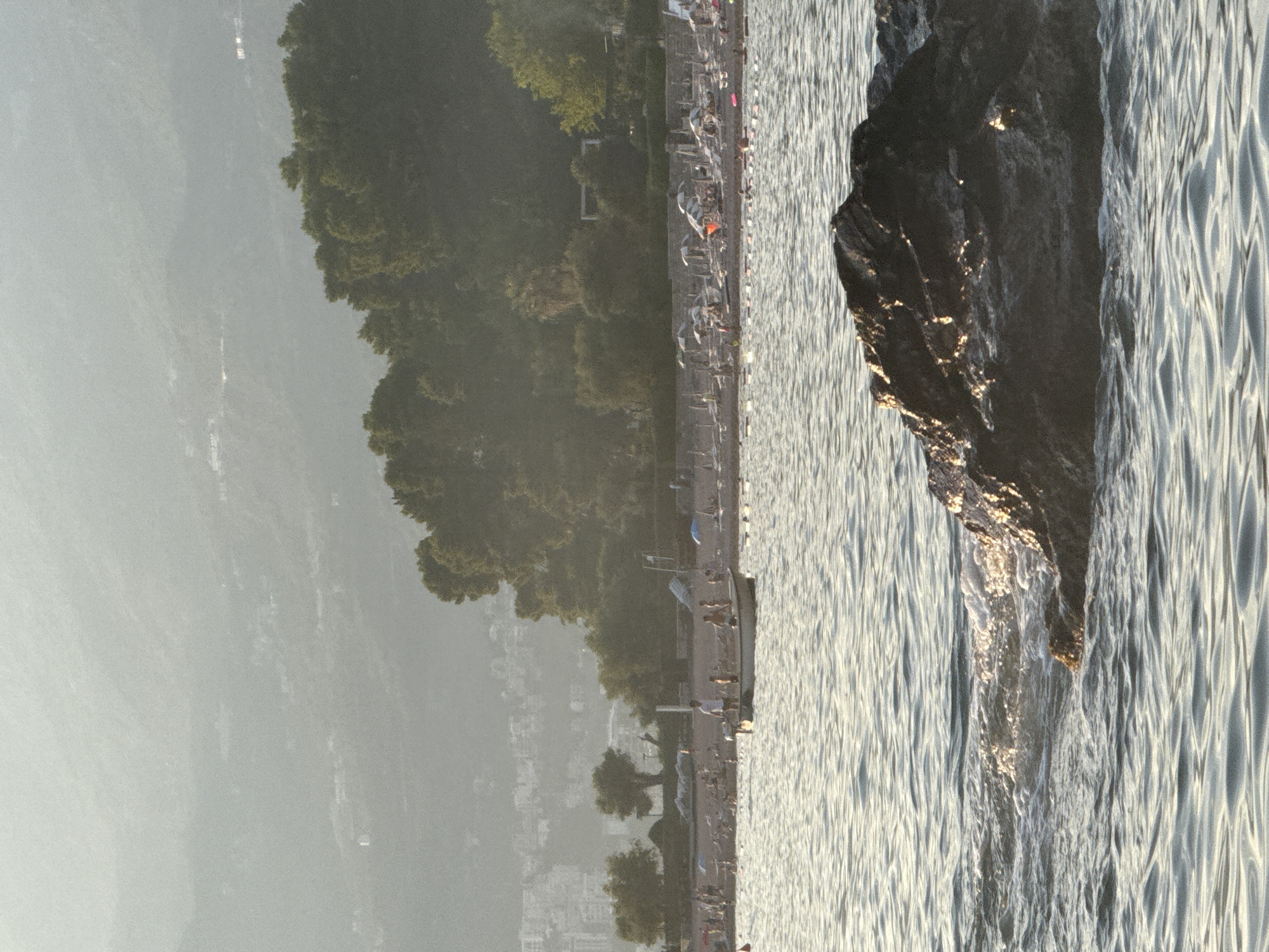 The image shows a rocky coastline with the sea on the right and a tree-lined shore on the left. The sun appears to be low in the sky, casting a warm glow on the water and the rocks. The sea is calm, with gentle waves lapping against the shore. The trees on the left are lush and green, providing a contrast to the ruggedness of the rocks. In the background, there are buildings and what appears to be a beach with people, suggesting a populated area. The atmosphere seems peaceful and serene.