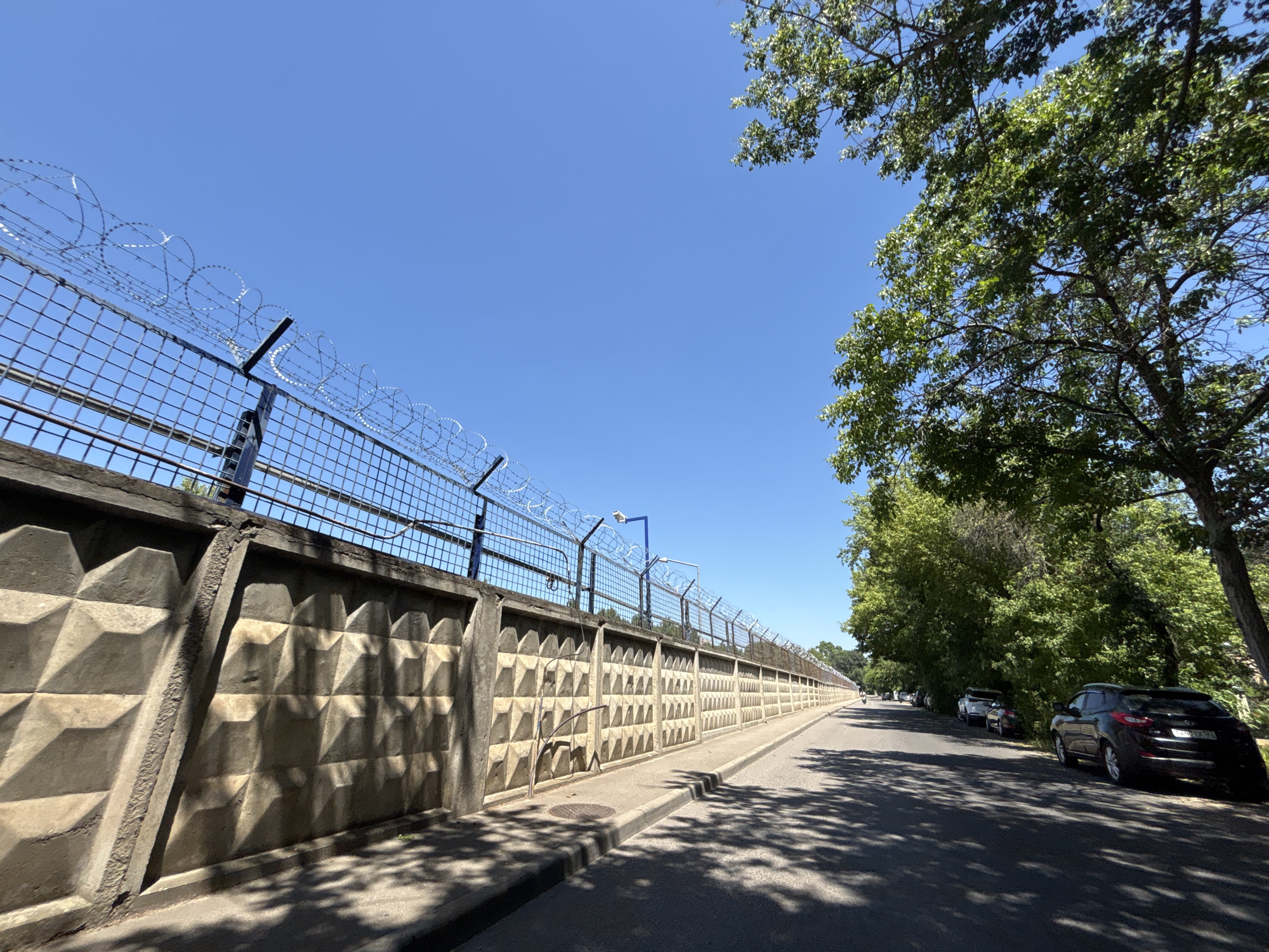 The image captures a sunlit outdoor scene, likely in Krasnodar, Russia, during the daytime under a clear blue sky. The focal point is a long, high concrete wall topped with a wire fence and razor wire, suggesting a secure or restricted area. The wall itself has a distinctive geometric, textured pattern.
To the left of the wall, a sidewalk runs alongside it, and further left, the wall begins. To the right of the wall, a paved road stretches into the distance, lined with parked cars. Lush green trees with dense foliage cast dappled shadows across the road and sidewalk, indicating the presence of abundant vegetation. The overall mood is tranquil and ordinary, with no specific event or activity prominently displayed.
The time of day appears to be mid-morning to early afternoon, given the bright sunlight and the distinct shadows. The weather is fair and warm, suitable for outdoor activities. While no people are actively engaged in significant actions, a person can be seen on a motorcycle in the distance on the right side of the road, and other parked cars suggest potential activity beyond the immediate frame. No visible text is present in the image.