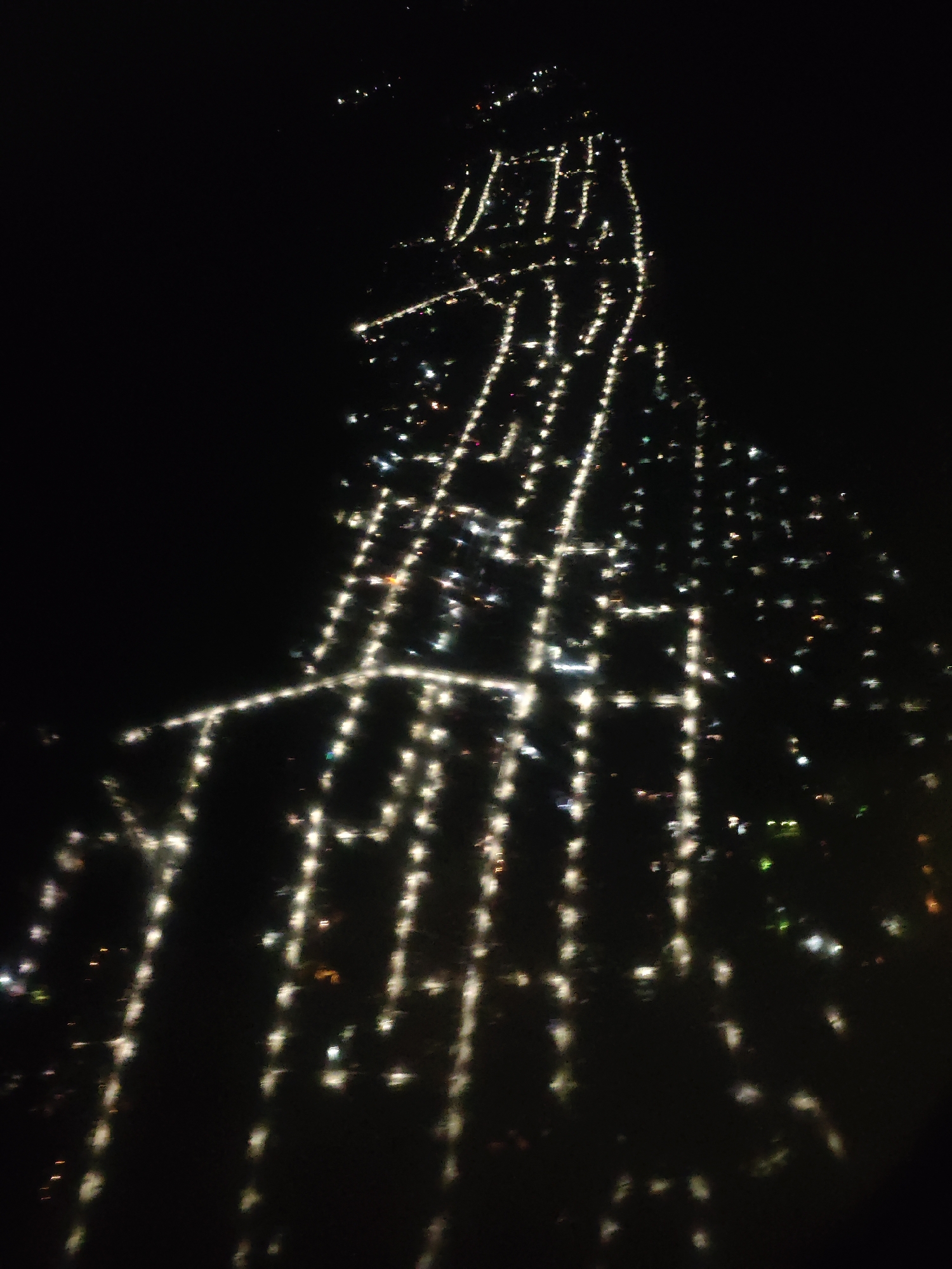 The image depicts a night view of Novosibirsk, Russia, from an elevated perspective, possibly from an airplane. The scene is dominated by streetlights forming a grid-like pattern, illuminating streets below. The lights appear slightly blurred due to the moving camera. The environment is dark, and the city lights are the primary source of illumination.