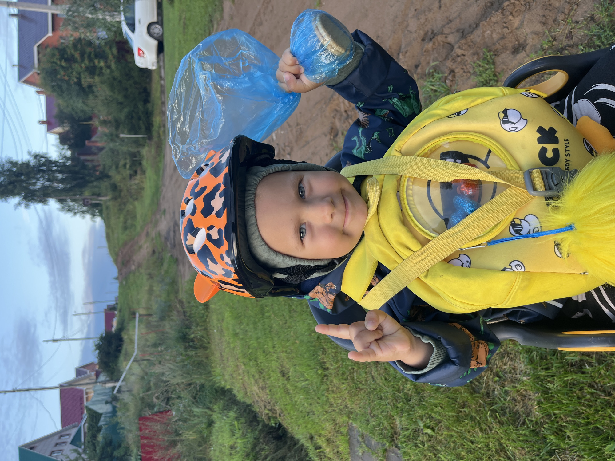 A young boy wearing a blue jacket with a dinosaur print and a yellow backpack with duck characters, sits in a stroller. He has on an orange and black leopard-print helmet with ear-like protrusions. In his right hand, he holds a blue plastic bag containing an object, and his left hand is raised in a "rock on" gesture. He is outdoors, with a dirt road behind him and grassy slopes and houses visible in the background. The sky is overcast with a hint of blue. The setting appears to be a residential area with rural characteristics, consistent with the outskirts of Chaikovsky, Russia.
