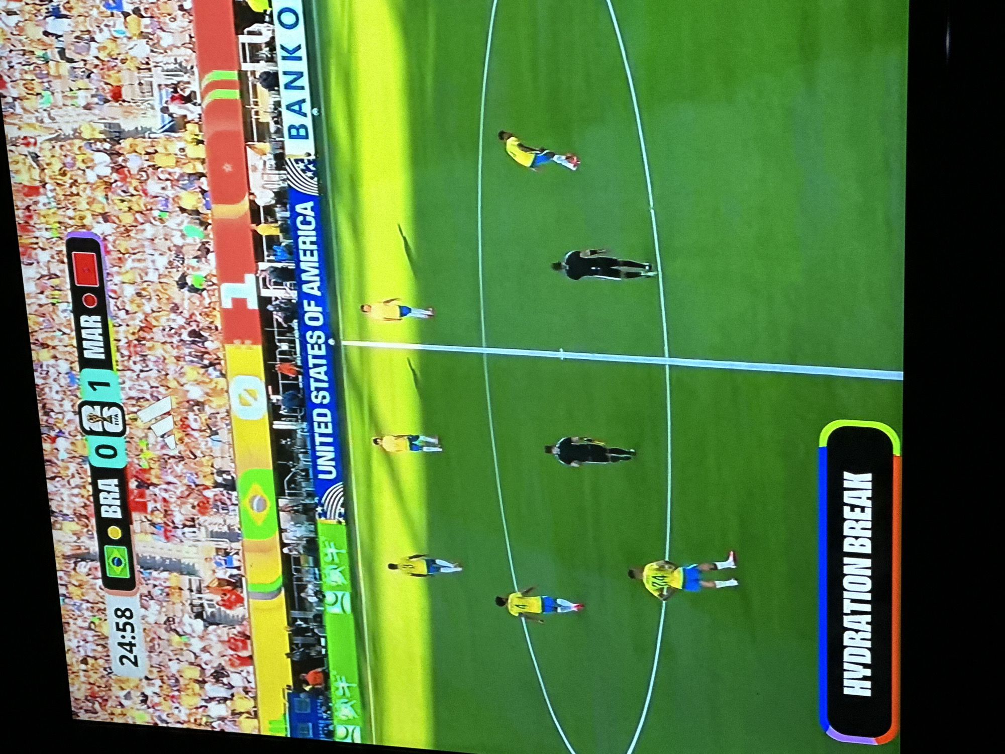 The image shows a soccer match in progress, with the score displayed as Brazil 0, Morocco 1. The time is 24:58. There are several players on the field, some in yellow jerseys and blue shorts, and one in a black and white referee uniform. The field is green grass, with white lines marking the boundaries and center circle. In the background, a stadium filled with spectators is visible, many wearing yellow. A banner above the spectators reads "UNITED STATES OF AMERICA BANK O". A prominent "HYDRATION BREAK" graphic with colorful borders is displayed in the lower portion of the image. The scene is captured on a screen, likely a television or monitor, and the image quality suggests it was taken from a distance. The specific location within Gura Top, Nigeria is not identifiable from the visual information provided.