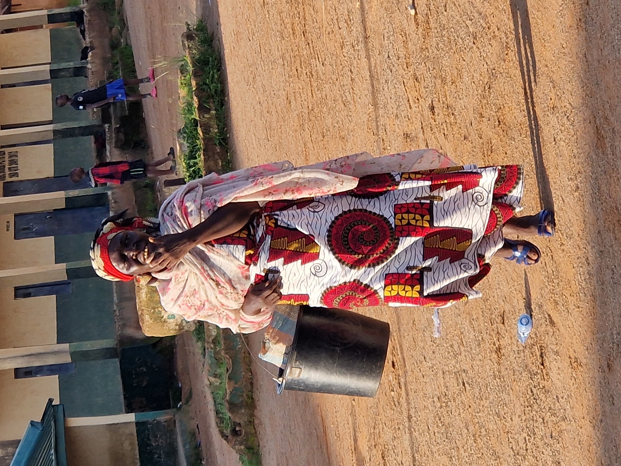 A medium shot shows a woman in a vibrant, patterned dress and a headscarf walking towards the camera in Kabong, Nigeria. She holds a black bucket in her left hand. The woman has her right hand near her mouth. In the background, two young boys stand in front of a building with a green stripe along its base. The ground is dirt and sand, with some patches of grass. A plastic water bottle lies on the ground in the foreground. The lighting suggests it is daytime.
