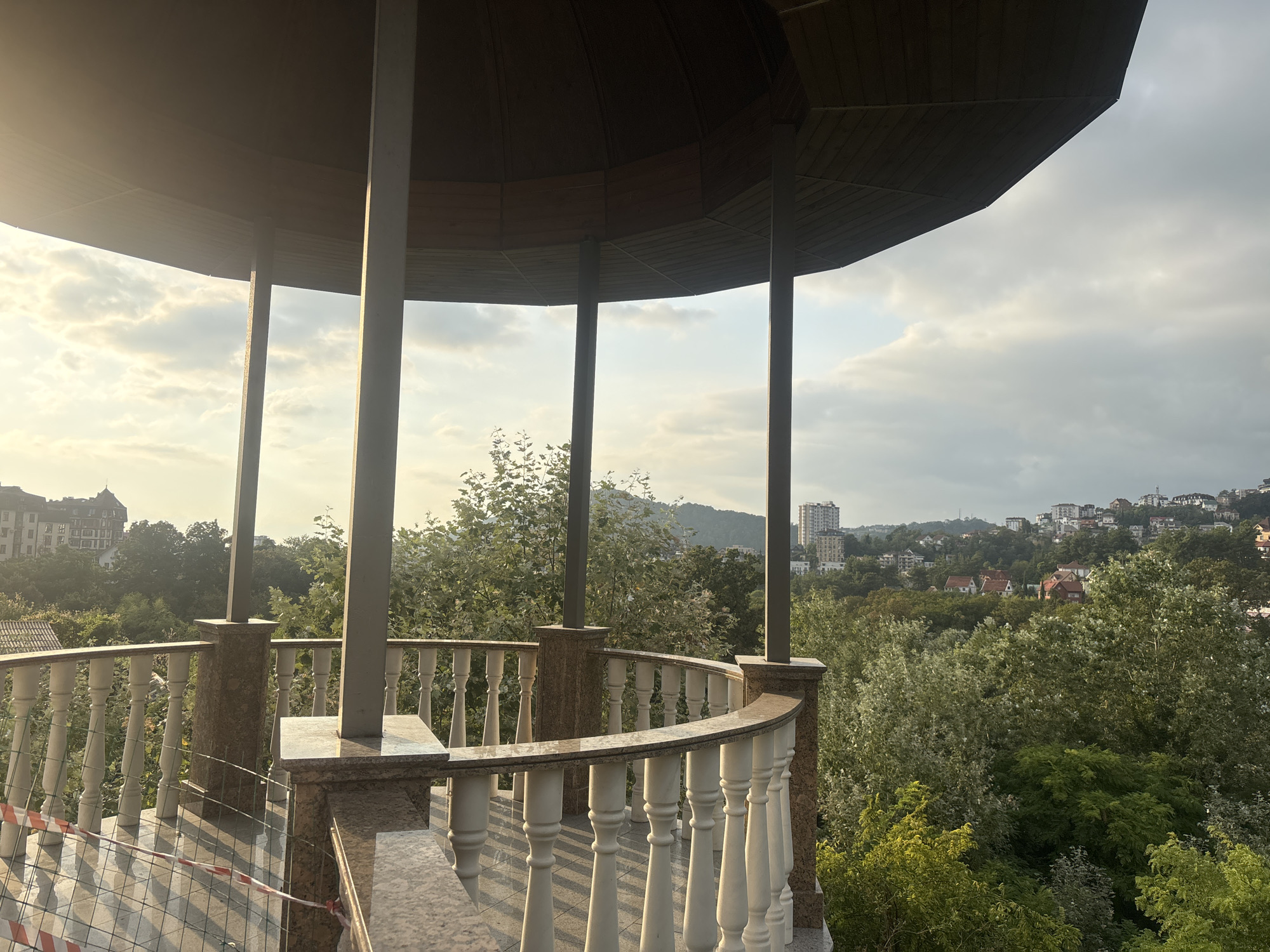 The image depicts a gazebo with a rounded balustrade, overlooking a verdant landscape with buildings in the distance. The gazebo's roof is made of dark wood panels, supported by four pillars. The balustrade is made of white balusters and a marble railing. The ground inside the gazebo is tiled and there is construction fencing with red and white tape. The background shows a dense forest with various shades of green foliage, and beyond that, a city with buildings of different sizes and architectural styles, including apartment blocks and houses, situated on hills. The sky is partly cloudy with the sun casting a warm glow, suggesting it is either early morning or late afternoon. The architecture and the presence of tall apartment buildings on the hills are characteristic of the city of Sochi, Russia, particularly areas overlooking the Black Sea coast.