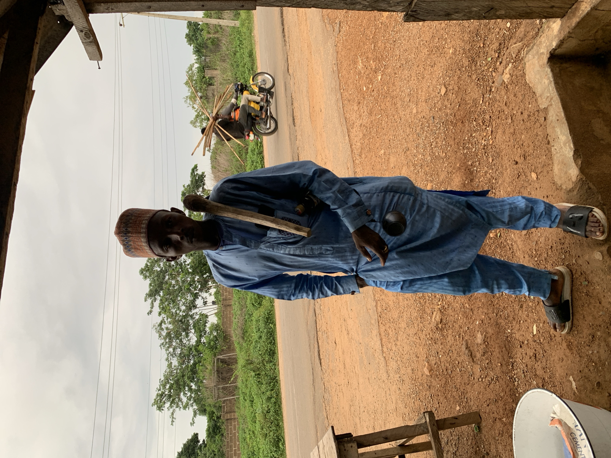 This image depicts a rural scene in Gbeleko, Nigeria, centered on a man standing under a wooden structure. The man, with dark skin, stands facing forward, wearing a light blue traditional tunic and trousers with a patterned reddish-brown cap. A long wooden stick rests over his left shoulder, and he holds a small, dark, gourd-like object in his right hand. His left hand is placed in his pocket. He wears black and white slide sandals. To his left, a white metal bucket containing a partial bag, possibly cement, is visible next to a crude wooden table. The ground is reddish-brown dirt and gravel.
In the midground, a paved road stretches horizontally, separating the foreground from a strip of green vegetation and a low, reddish-brick wall. Behind this, more dense green foliage and trees are visible. A yellow motorcycle with two occupants travels from left to right on the road. The passenger on the motorcycle carries a large, fanned bundle of thin wooden sticks on their back. The rider wears dark clothing, and the passenger appears to wear an orange vest. The sky is overcast with power lines traversing the upper portion of the frame. To the far right, a rough wooden beam frames the scene, indicating the photographer's position under a rudimentary shelter.
