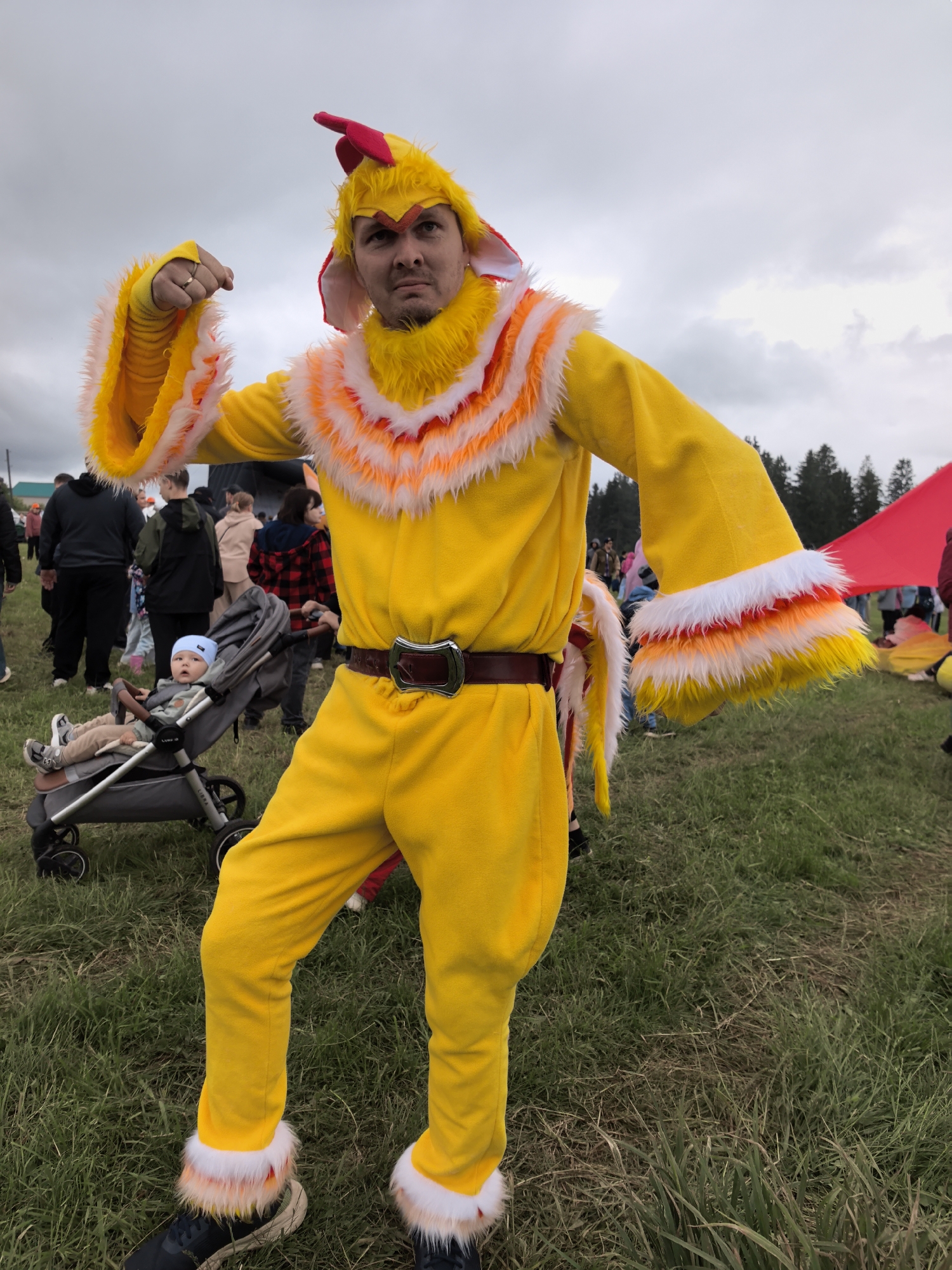 A male individual, approximately 30-40 years old, stands centrally in a full-body bright yellow chicken costume outdoors. The costume features a red comb headpiece, floppy ear-like appendages, a feathered collar and cuffs in white, orange, and yellow, and a brown belt with a silver buckle. He flexes his left bicep, displaying a silver ring on his left ring finger, and has a serious or slightly stern expression directed at the viewer. His footwear consists of dark sneakers, partially visible beneath the costume's ankle fringe. To his left, a baby wearing a light blue hat is seated in a grey stroller, looking forward. In the background, numerous out-of-focus individuals are gathered on a grassy field. A line of dark green conifer trees is visible in the distant background, and a large red fabric object, possibly a banner or tent, is present on the right mid-ground. The sky is overcast with grey clouds. The ground is covered in green and dry grass. The scene is located in Unknown, Unknown.