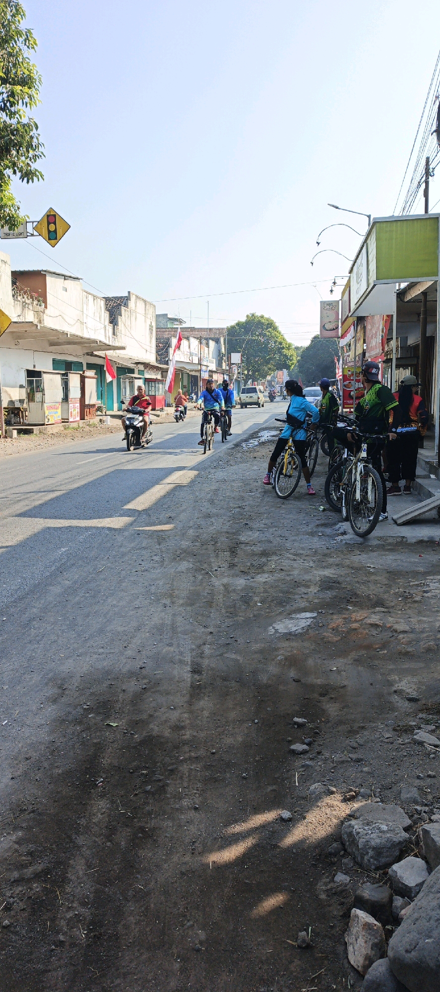 This is a photograph of a street scene in Menampu, Indonesia. Several people are gathered on the side of a road with bicycles. One person on a motorcycle is in the middle of the road. Buildings line the street, and Indonesian flags are visible. The sky is clear and blue, suggesting it is daytime. The ground is unpaved and dusty, with some scattered rocks and debris. There are shadows cast on the road, indicating the sun is at an angle.
