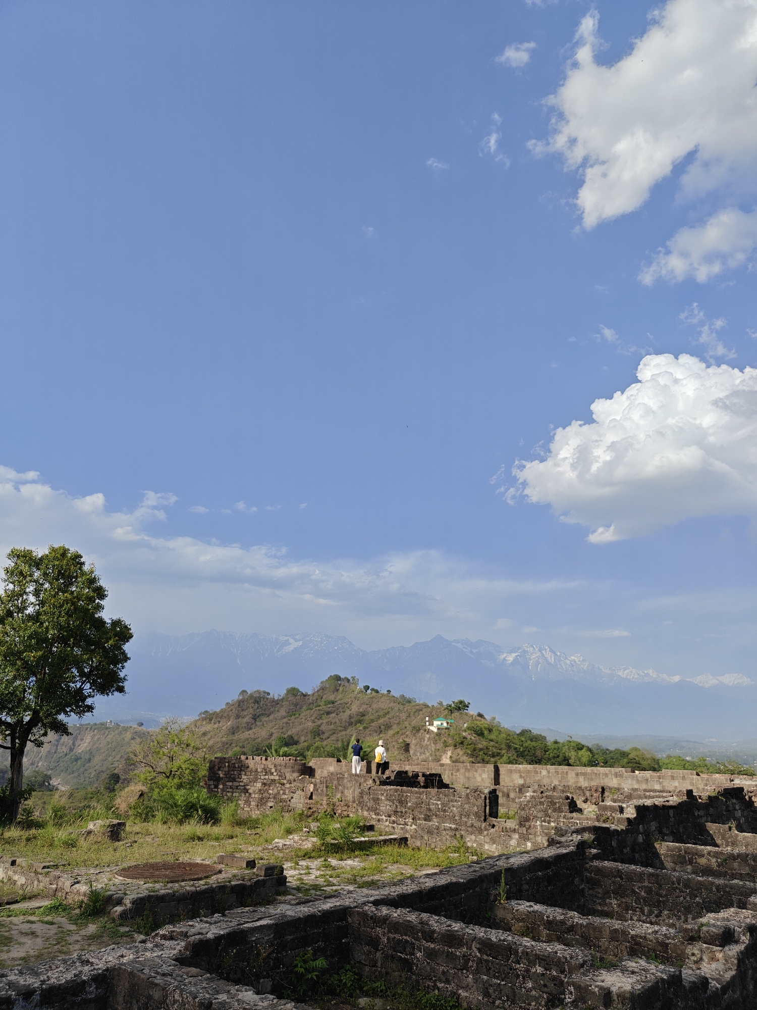 The image depicts the Kangra Fort ruins near Kaswara, India, under a blue sky with scattered clouds. In the foreground, crumbling stone walls and foundations of the fort are overgrown with grass and weeds. A metal grate is visible in the middle ground. Two individuals stand atop a section of the fort's wall, appearing to observe the scenery. Behind them, a sloping hillside is covered with trees, and in the distance, snow-capped mountain peaks are visible. A solitary tree stands on the left side of the image.