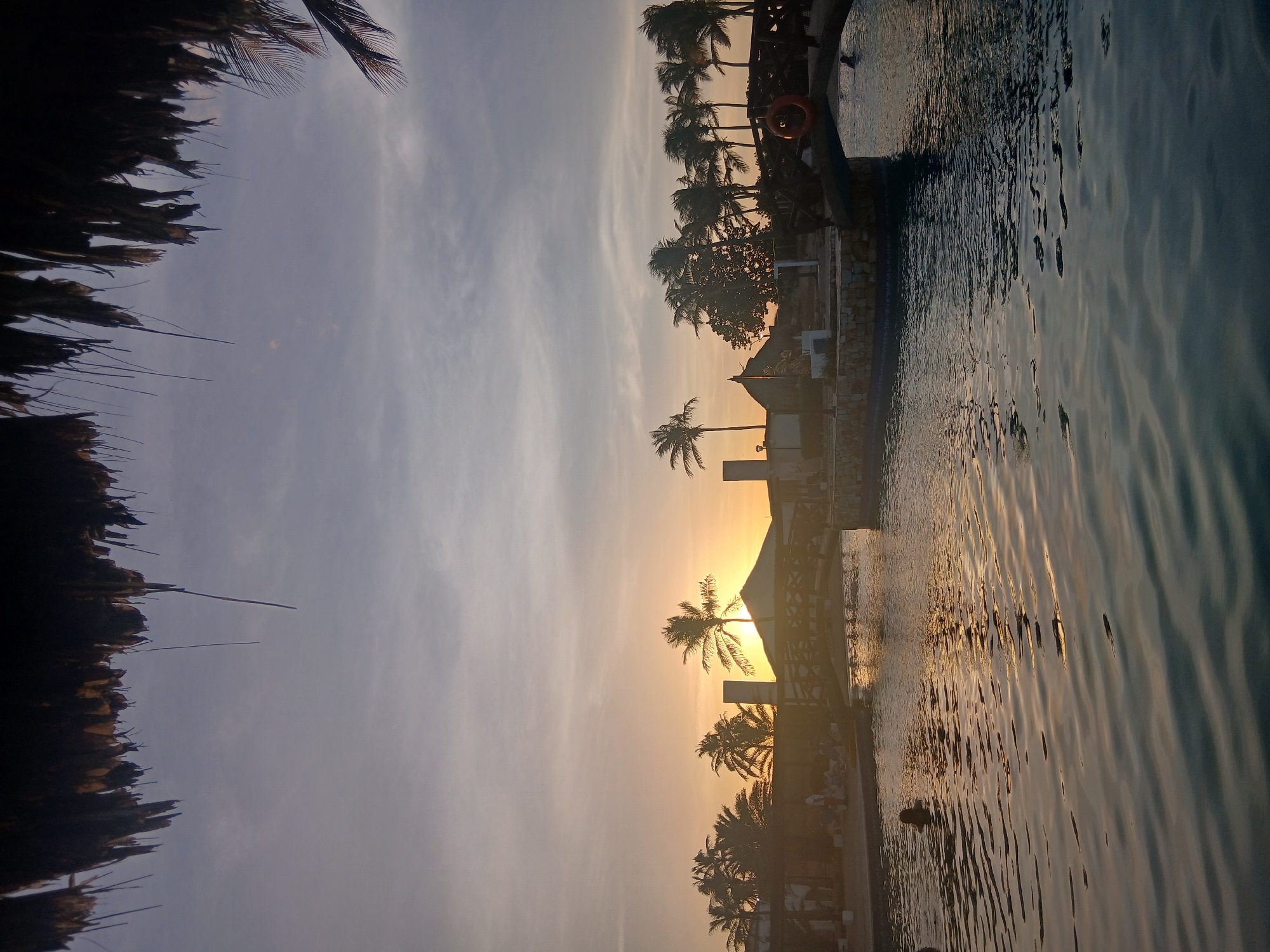 This image depicts an outdoor resort scene in Altagracia, Venezuela, during either sunrise or sunset. The perspective is from beneath a dark, out-of-focus thatched roof, with fragments of palm fronds visible in the upper frame. The sky exhibits an orange and yellow glow at the horizon, transitioning to a light blue-grey overhead with faint stratocumulus clouds. The sun is low on the horizon, centrally positioned behind a building with a peaked, possibly thatched, roof and several palm trees, creating strong backlighting and silhouettes.
A large body of water, consistent with a swimming pool, occupies the lower half of the image, displaying ripples and reflecting the sky's warm light. The dark silhouette of an individual's head is visible in the water on the lower-left. A dark wooden bridge with an arched underside spans a section of the pool, extending from the right towards the center. A red life preserver ring is suspended from the bridge's railing on the right side. On the far side of the pool, multiple individuals are visible in silhouette on a paved deck area, some appearing seated or lounging. Numerous palm trees and additional resort structures with peaked roofs are discernible beyond the pool, further silhouetted against the bright sky.