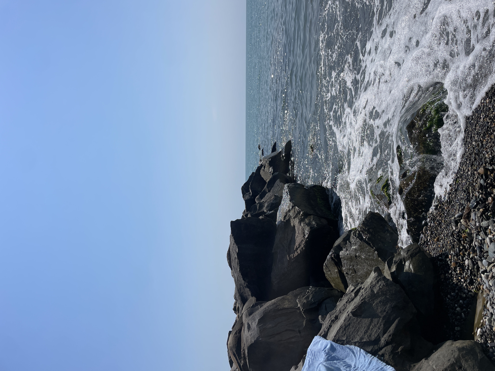The image depicts a rocky shoreline on a clear, sunny day, likely in Sochi, Russia, given the context. The scene is dominated by large, dark gray rocks that form a breakwater, with smaller pebbles and stones lining the shore in the foreground. Gentle waves crash against the rocks, creating white foam and splashing water. The sea is a deep blue, shimmering with sunlight. A light blue towel is draped over one of the rocks on the left side of the image. The sky is a clear, pale blue, with no clouds visible. The overall impression is one of a peaceful, natural coastal environment.