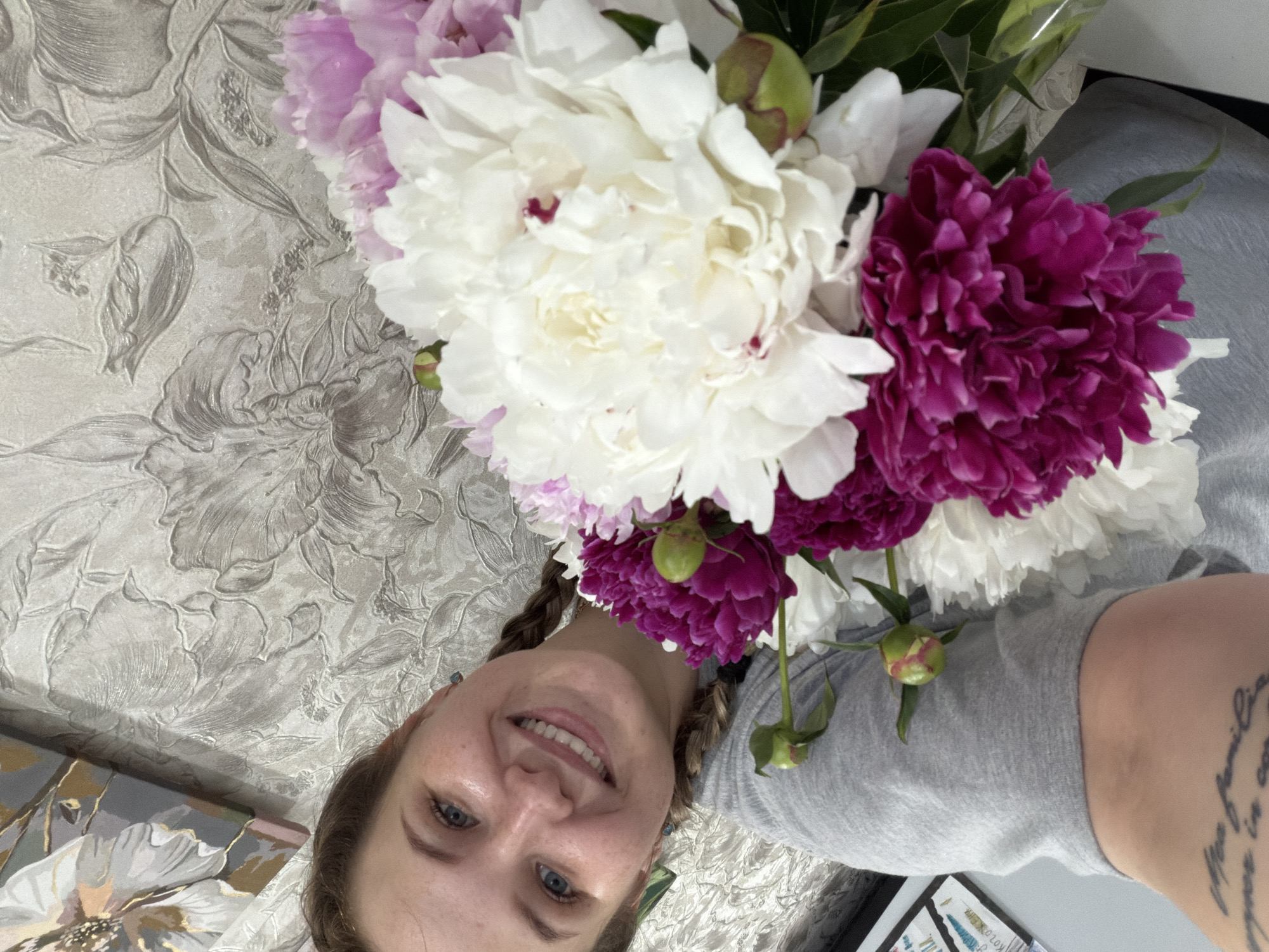 A young woman with blue eyes, freckles, and braided hair smiles at the camera while holding a bouquet of white and pink peonies. She is wearing a gray t-shirt. The background consists of floral-patterned wallpaper. A painting with abstract floral designs hangs on the wall above and to the left of the woman. The scene appears to be indoors, likely in a residential setting in Chaikovsky, Russia.