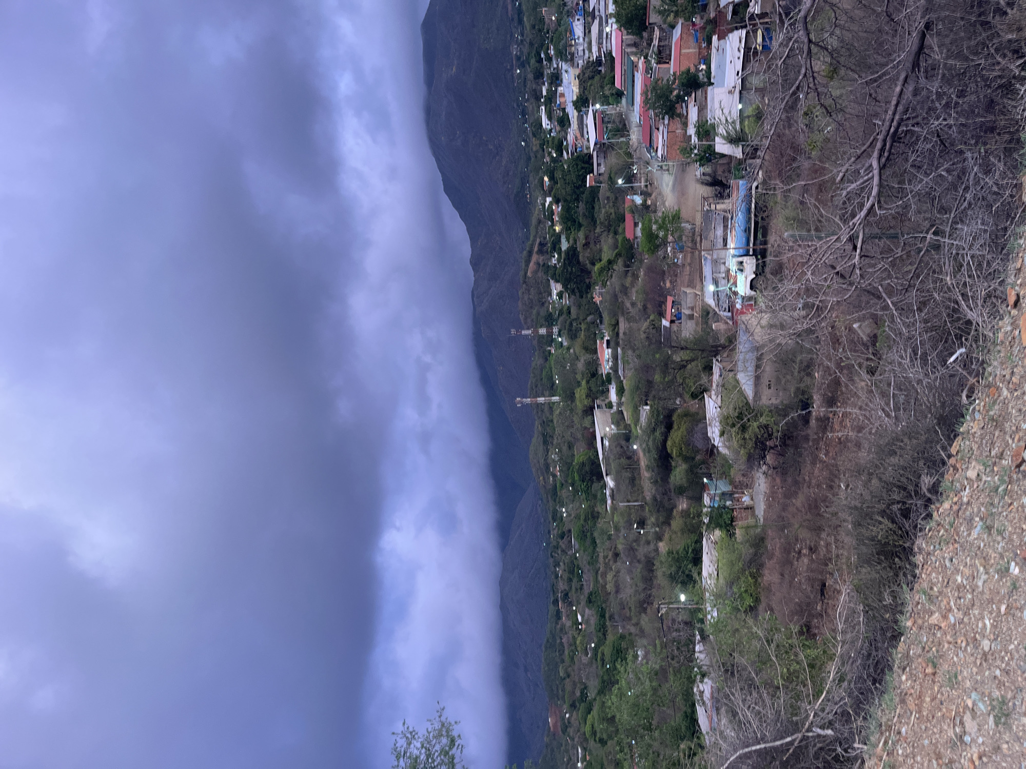 The image shows a view of a mountainous landscape with a town nestled in the valley. The sky is overcast with dark, heavy clouds, suggesting an impending storm. In the foreground, there is dry, brushy vegetation and rocky terrain. The middle ground reveals a cluster of buildings with red and gray roofs, characteristic of a town or village. Some of these buildings appear to be made of brick and concrete. A white truck with a blue tarp-covered cargo is visible on a dirt road within the town. In the background, rolling hills and larger mountains are shrouded by low-lying clouds. A telecommunications tower stands prominently on one of the hills. The overall scene depicts a rural or semi-urban environment in the city of Santa Ana, Venezuela, under a dramatic sky.