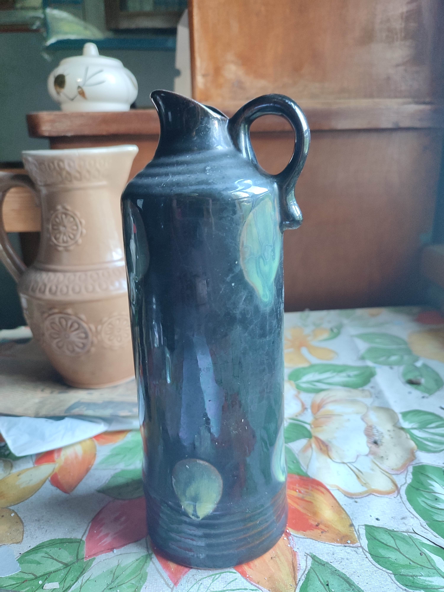 The image shows a still life composition of pottery objects placed on a floral-patterned tablecloth. The foreground is dominated by a dark, glossy ceramic jug with a curved handle and a spouted opening. This jug features irregular, light green circular accents. To the left and slightly behind, a larger, lighter brown ceramic pitcher with a prominent handle is visible. This pitcher has embossed circular decorative patterns around its body. In the background, a white ceramic lidded pot with painted details rests on a wooden surface. The overall scene appears to be indoors, with a wooden structure forming the backdrop. The image is static and captures these objects from a slightly elevated, frontal perspective. There is no indication of Gremyachinsk, Russia in the image content.