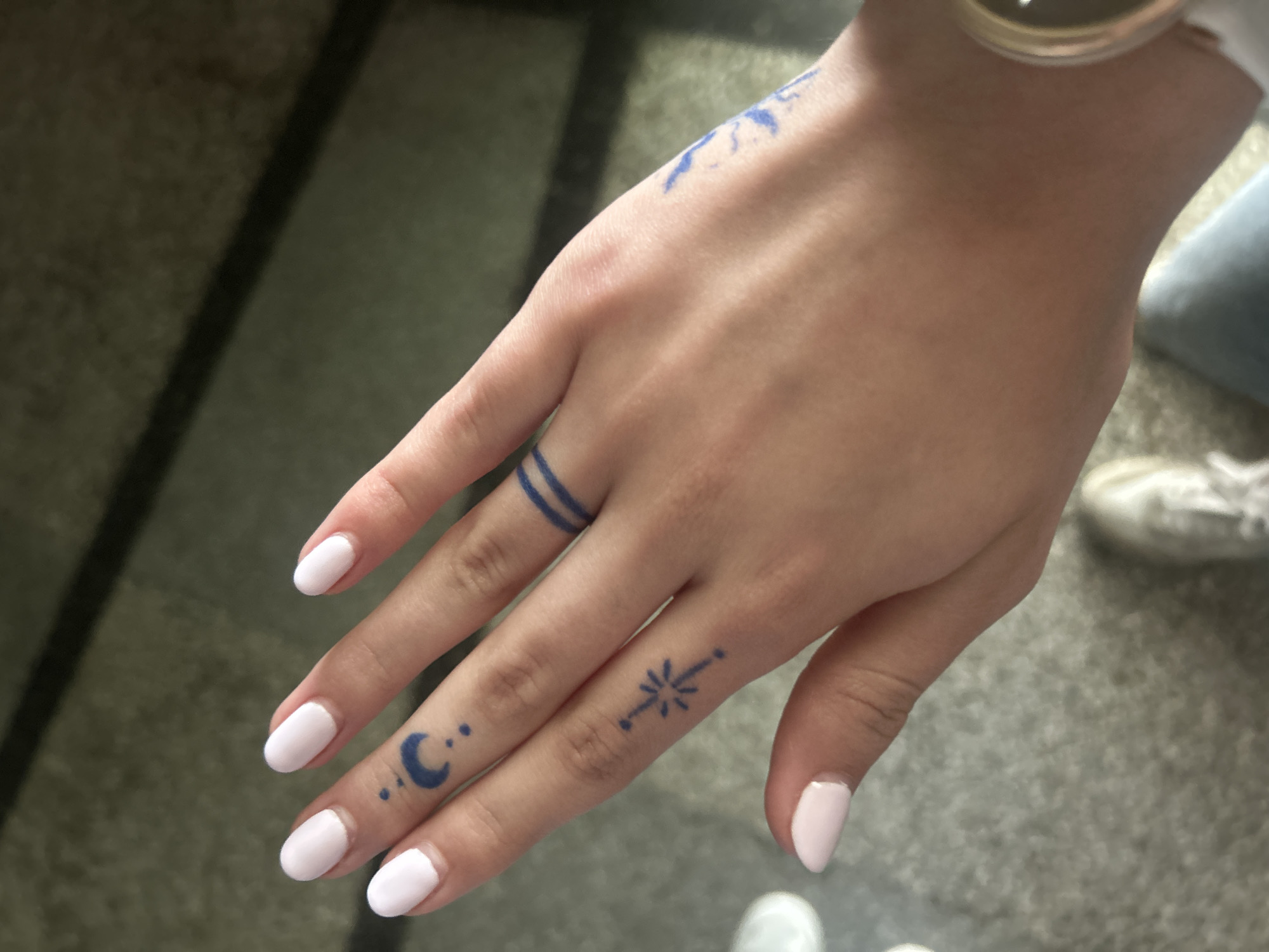 The image depicts a fair-skinned hand with manicured nails and several blue tattoos, including two parallel lines on one finger, a crescent moon and dots on another, and a starburst-like design on a third finger. An additional tattoo design is partially visible on the back of the hand. The background appears to be an indoor space with tile or linoleum flooring and partial views of clothing and a wristwatch. There is no indication of location in the city of Ruse, Bulgaria.