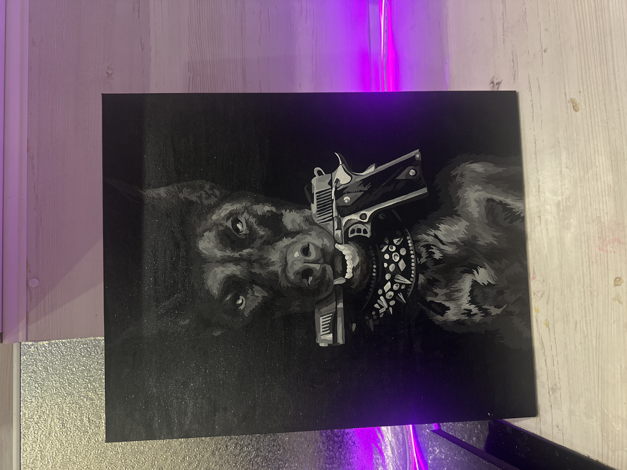 The image depicts a black and white painting of a dog holding a handgun in its mouth. The dog also wears a studded collar. The painting is placed against a wall with a light source casting a purple hue from the top. The location is unknown.