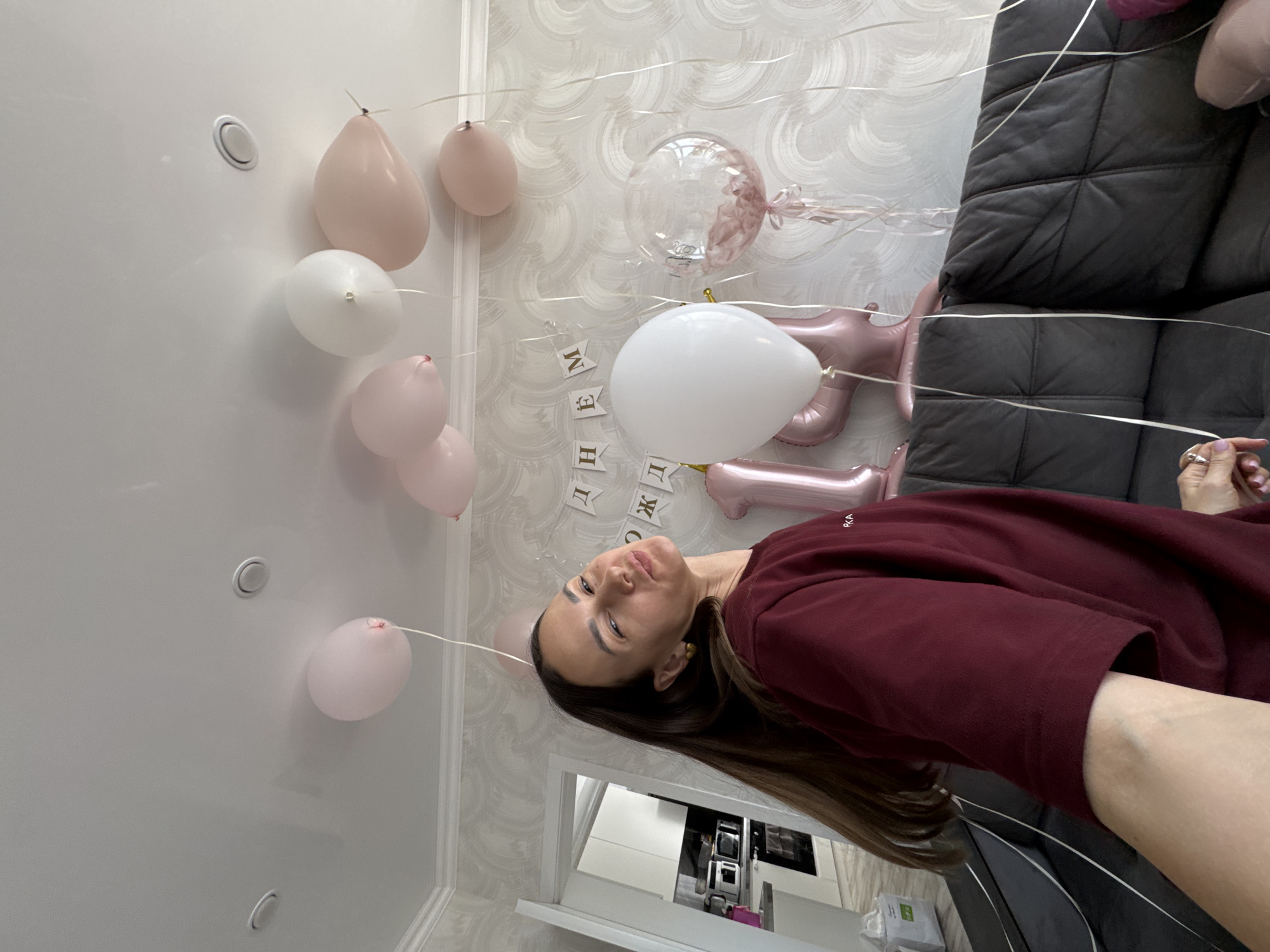 The image shows a woman standing in a room decorated for a birthday celebration. The room has wallpaper with a subtle, textured pattern. Numerous balloons in shades of white and pink are attached to the ceiling. Some of the balloons are shaped like the numbers '1' and '7', indicating the age of the birthday celebrant. There is also a banner with text in Russian. The woman is wearing a maroon shirt and is holding a white balloon. A dark grey couch is partially visible in the background. A glimpse of a kitchen can be seen through an open doorway, including appliances.
It's difficult to confirm the location is in Sochi, Russia solely from the visual details of the image.