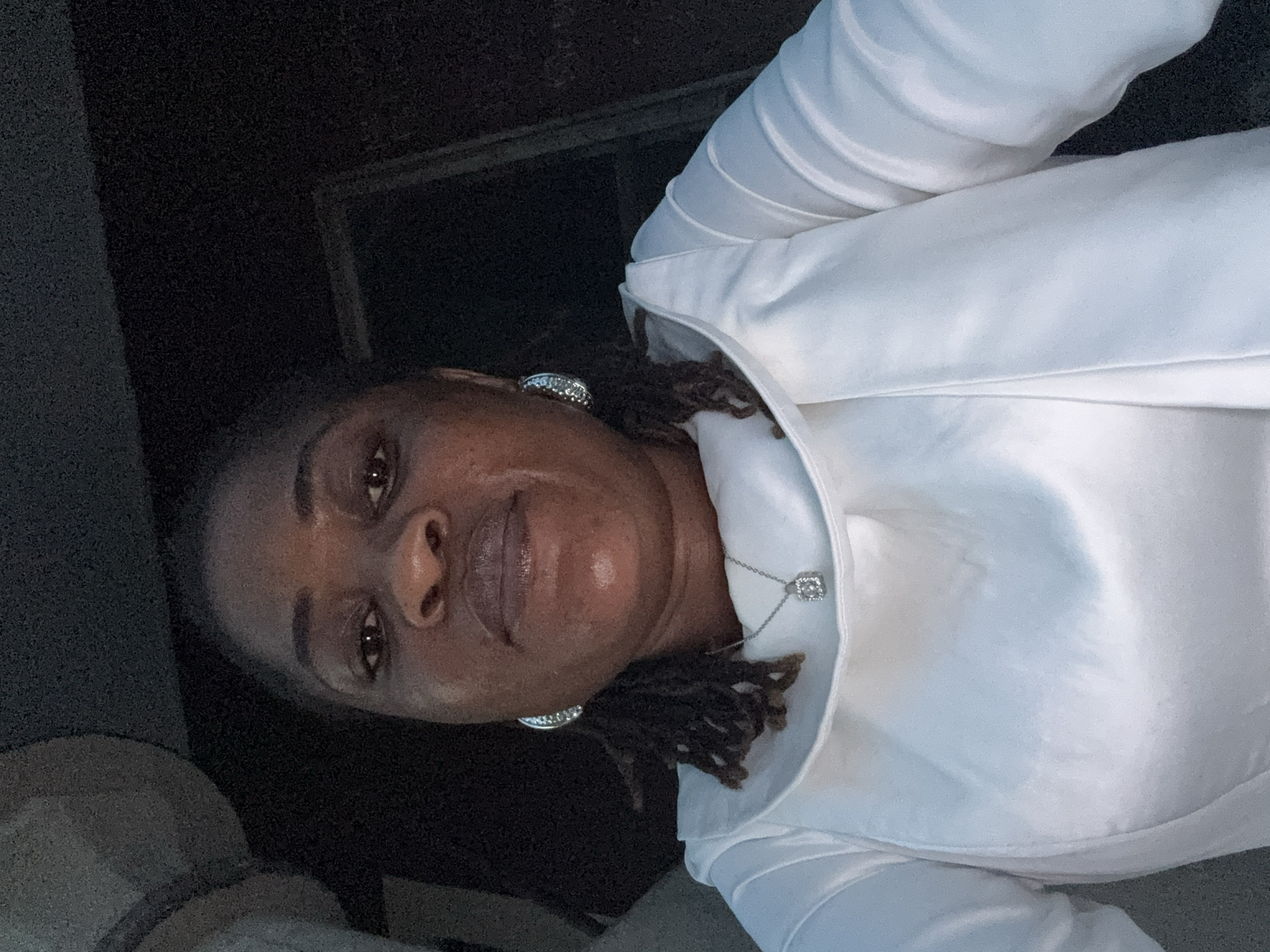 The image is a close-up selfie of a Black woman in Badagry, Nigeria. She is wearing a white blouse and a silver necklace with a square pendant. She has dark, braided hair and is wearing large, silver, hoop earrings. Her facial features are dark with brown eyes, a slightly broad nose, and full lips. The background is dark and appears to be an indoor setting, possibly a car, given the angle and lighting. The overall impression is that of a candid portrait.