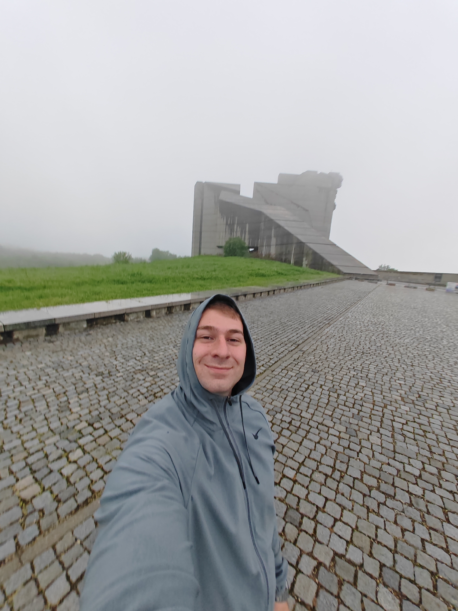 A man in a grey hooded jacket takes a selfie in front of the Monument to 1300 Years of Bulgaria, located in Shumen, Bulgaria. The monument is large and concrete, partially obscured by fog. The man is smiling. The ground is paved with cobblestones, and there is a grassy area to the left. The weather appears foggy.