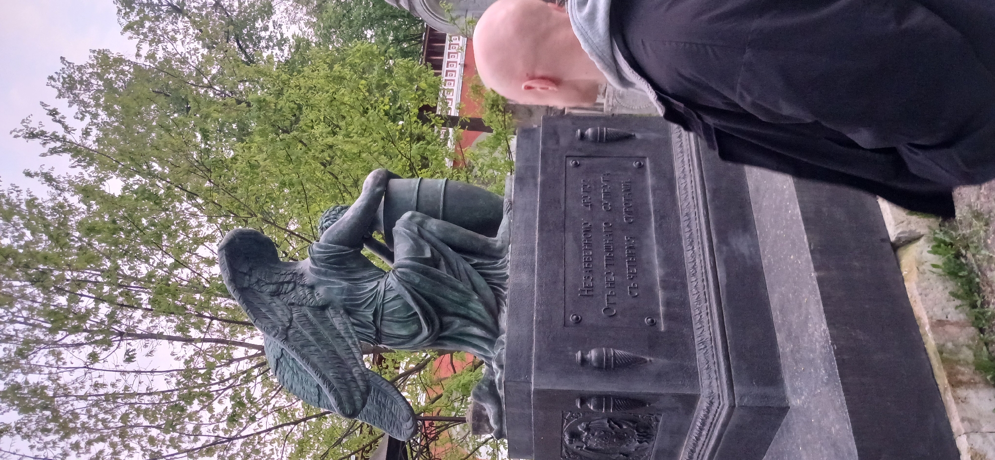 Here is a description of the image:
The image shows a bronze statue of a weeping angel leaning against a funerary urn atop a dark, stepped monument. The statue and monument are likely in a cemetery. A bald-headed person wearing a dark jacket is leaning over the monument, possibly reading the inscription. In the background, trees with green leaves are visible. Based on the inscription which contains Cyrillic characters, the monument is located in the city of Moscow, Russia.