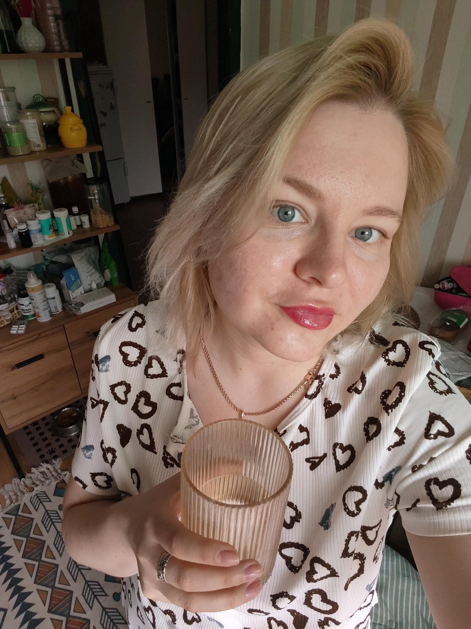 A woman with blonde hair and blue eyes is holding a glass of water in front of her in an indoor environment. She is wearing a white shirt with brown heart patterns and a gold necklace. A silver ring is visible on her finger. Behind her is a wooden shelf filled with jars, bottles, and other items, suggesting a kitchen or storage area. A white refrigerator is also visible in the background, along with vertically striped wallpaper on a wall. A rug is placed at the bottom of the image. There is no information in the image that the location is in Izhevsk, Russia.