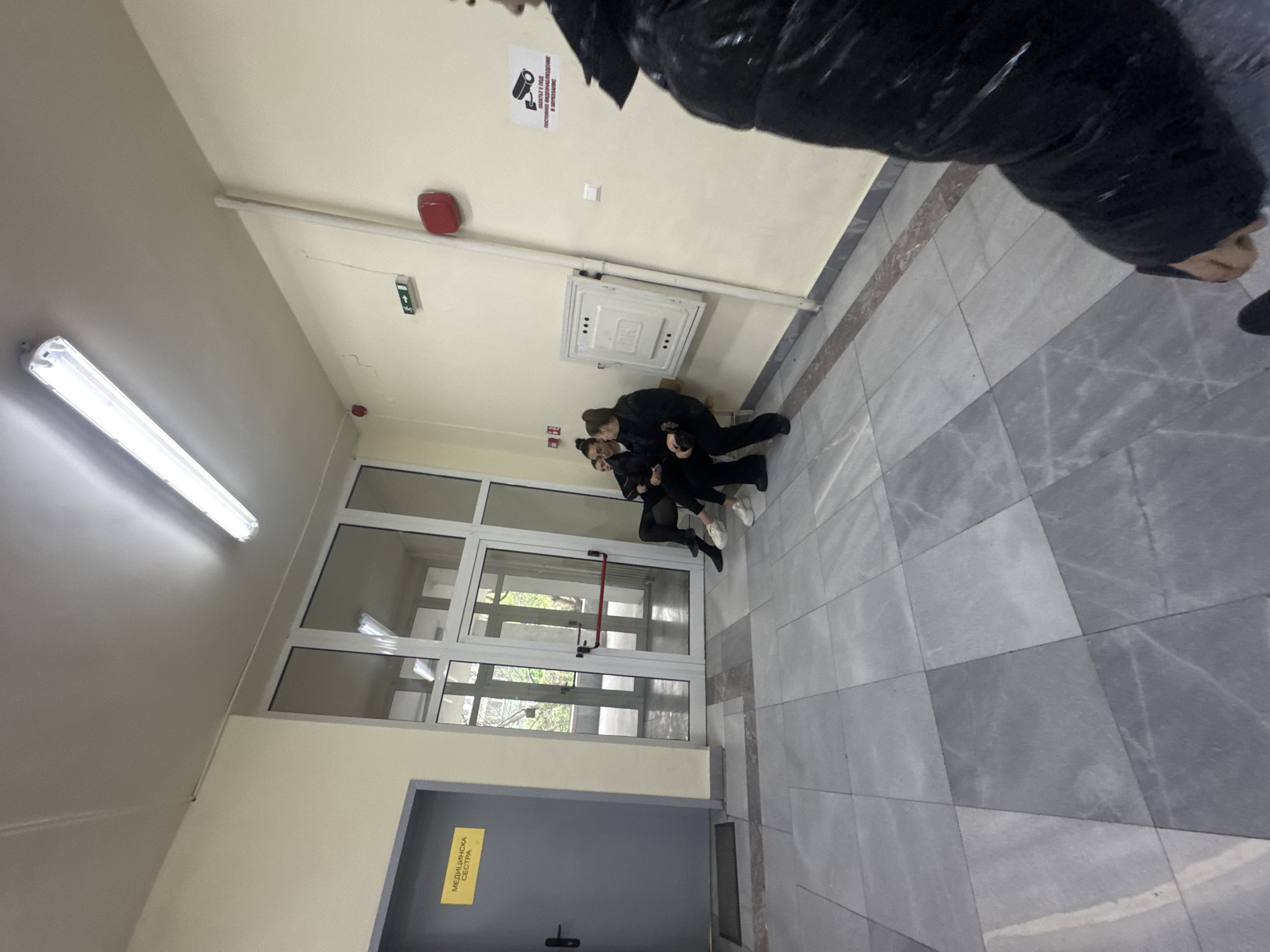 The image depicts an indoor hallway, likely within a public or institutional building, viewed from a slightly low angle. In the immediate foreground, part of a person's torso and arm, clad in a shiny black puffy jacket, is visible in the upper right corner, suggesting the photo was taken while walking or standing.
Further down the hallway, two individuals, appearing to be young women or teenagers, are seated on the tiled floor against a wall next to a large glass door and window partition. They are both dressed in dark clothing. One person, with hair tied back, is leaning against the wall, while the other, also with dark hair pulled up, is sitting with legs extended, holding what appears to be a smartphone. They seem to be resting or waiting.
The hallway features light-colored walls and a tiled floor composed of various shades of gray, creating a geometric pattern. On the ceiling, a long fluorescent light fixture illuminates the space. Other ceiling or upper wall fixtures include a red fire alarm pull station, a small white square panel, and a green sign with a white cross, indicating a first aid point. A larger white square access panel is also visible on the wall above where the two people are sitting.
A sign affixed to the wall near the top reads in Bulgarian: "ОБЕКТЪТ Е ПОД ПОСТОЯННО ВИДЕОНАБЛЮДЕНИЕ И ВИДЕОЗАПИС," which translates to "The object is under constant video surveillance and video recording." Below the people sitting, a gray door is visible with a yellow sign on it, also in Bulgarian: "МЕДИЦИНСКА СЕСТРА," meaning "Medical Nurse" or "School Nurse." Through the large glass door and window partition, some outdoor greenery can be seen, hinting at the building's exterior. The scene appears to be well-lit by artificial light.