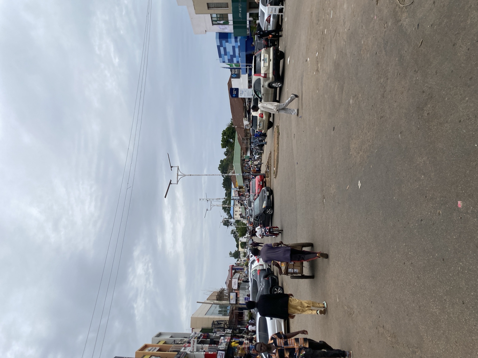The image shows a street scene in Jos, Nigeria, on an overcast day. The street is paved and appears to be busy with traffic and pedestrians. Several cars are parked on the side of the road, and others are driving. A man is pushing a handcart, and others are walking on the pavement. Buildings line both sides of the street, housing shops and businesses. The overall impression is of a bustling urban environment.