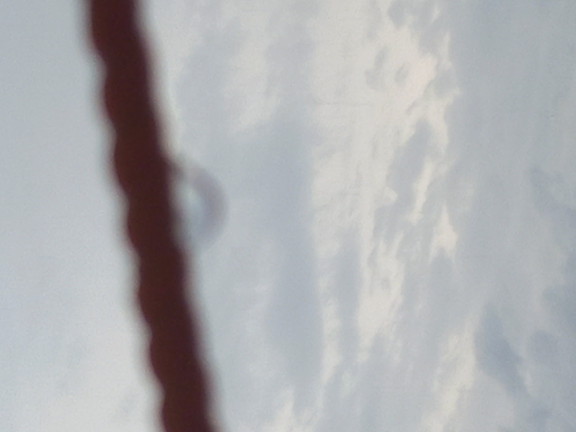 The media file presents an abstract, blurred composition dominated by a dark, vertical, elongated object in the foreground, likely a rope, cable, or similar structure, extending upwards and downwards out of the frame. This object is significantly out of focus, appearing soft and indistinct.
The background consists of a cloudy sky, rendered in varying shades of white and light grey-blue, suggesting a daytime scene with overcast weather. To the right of the dark foreground object, approximately in the upper-middle section of the sky, a very faint, blurred, and somewhat circular or semi-circular shape is discernible.
No people, discernible activities, or specific events are taking place. The image is static, focusing on environmental elements. There are no visible texts or specific location cues within the image itself to identify a particular place, though the provided context indicates Puloblang, Indonesia. The overall impression is one of soft focus, possibly an accidental shot or an artistic rendering emphasizing texture and light.