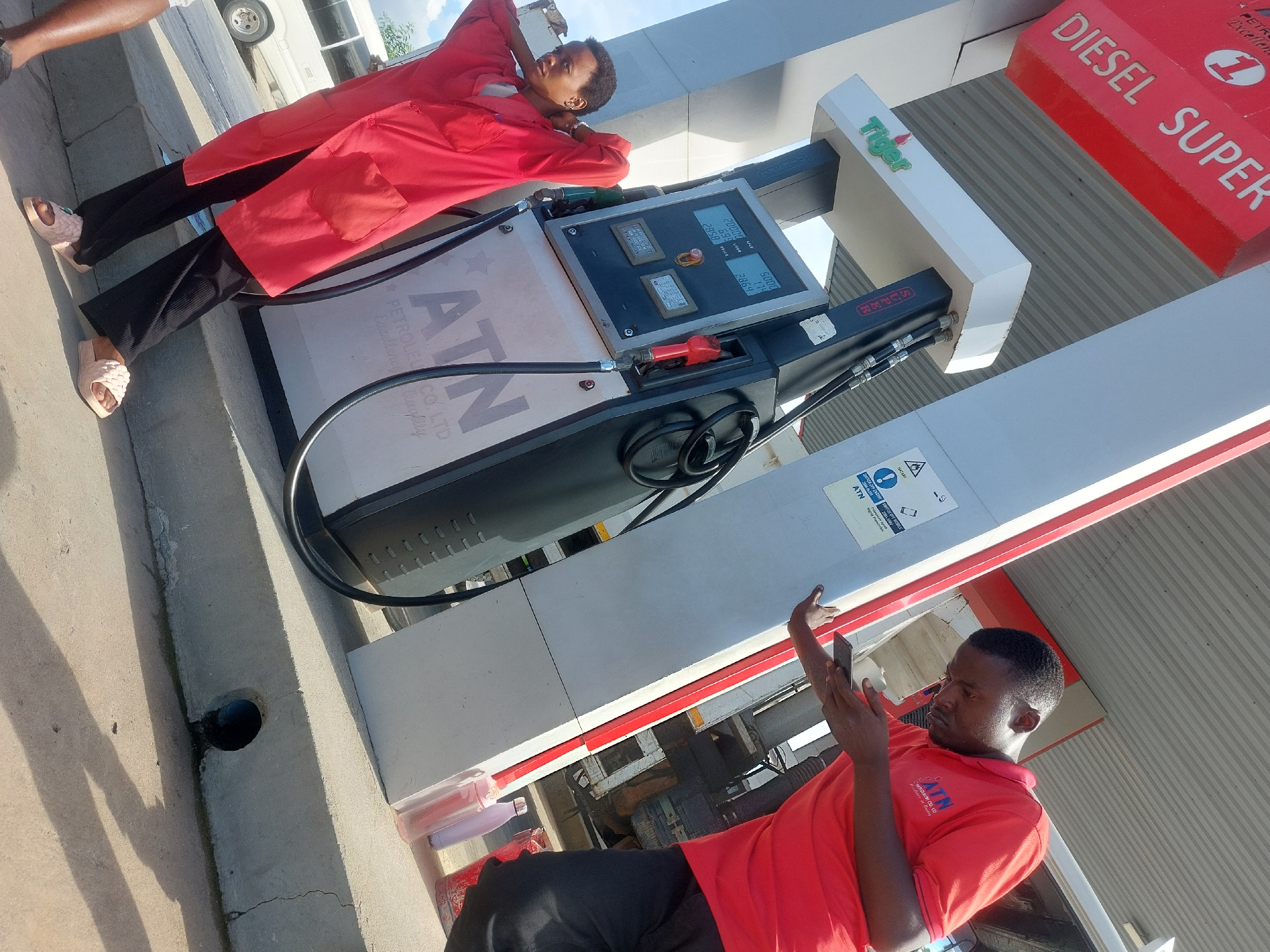 The image shows two people at an ATN gas station, seemingly in Dar es-Salaam, Tanzania. The scene is bright and sunny. One person, wearing a red uniform and black pants, is leaning against a gas pump, apparently resting. They are wearing pink sandals. Another person, also in a red uniform and black pants, is looking at a cell phone and is positioned near the gas pump, likely an employee. The gas pump is labeled "ATN" and has a display showing numbers, indicating a recent transaction. The station sells both Diesel and Super fuel. The environment is a typical gas station setting with a corrugated metal awning and a nearby vehicle.