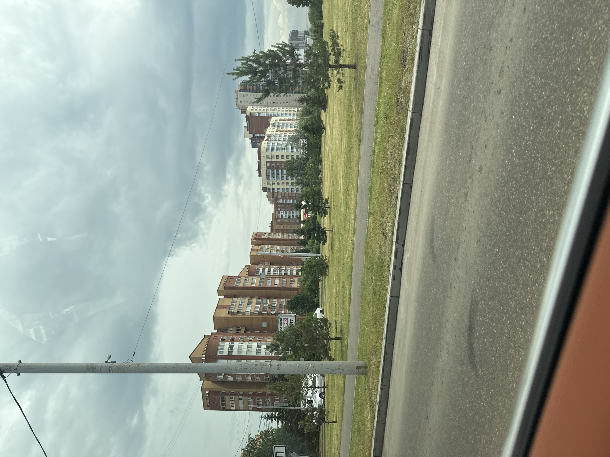 A wide shot captures a view from a moving vehicle looking towards a line of modern apartment buildings under a cloudy sky in Krasnoyarsk, Russia. The buildings, predominantly brick and light-colored, vary in height and architectural style, suggesting a developed urban area. In the foreground, a paved road and a grassy median with scattered young trees are visible. A tall, weathered utility pole stands prominently on the left side of the frame, equipped with a camera and wires. A few cars are parked or driving on the road, and a white sign with a black symbol is affixed to the utility pole. The overall scene depicts a typical urban streetscape on a day with overcast conditions.