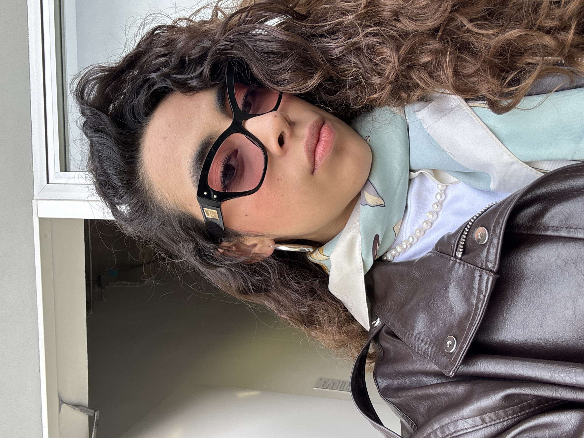 A woman with brown curly hair is wearing black-rimmed sunglasses with rose-colored lenses, a pearl necklace, and a brown leather jacket. A light blue scarf is visible around her neck. The image appears to be taken indoors near a window, but the exact location in Mexico City, Mexico cannot be determined without additional context.