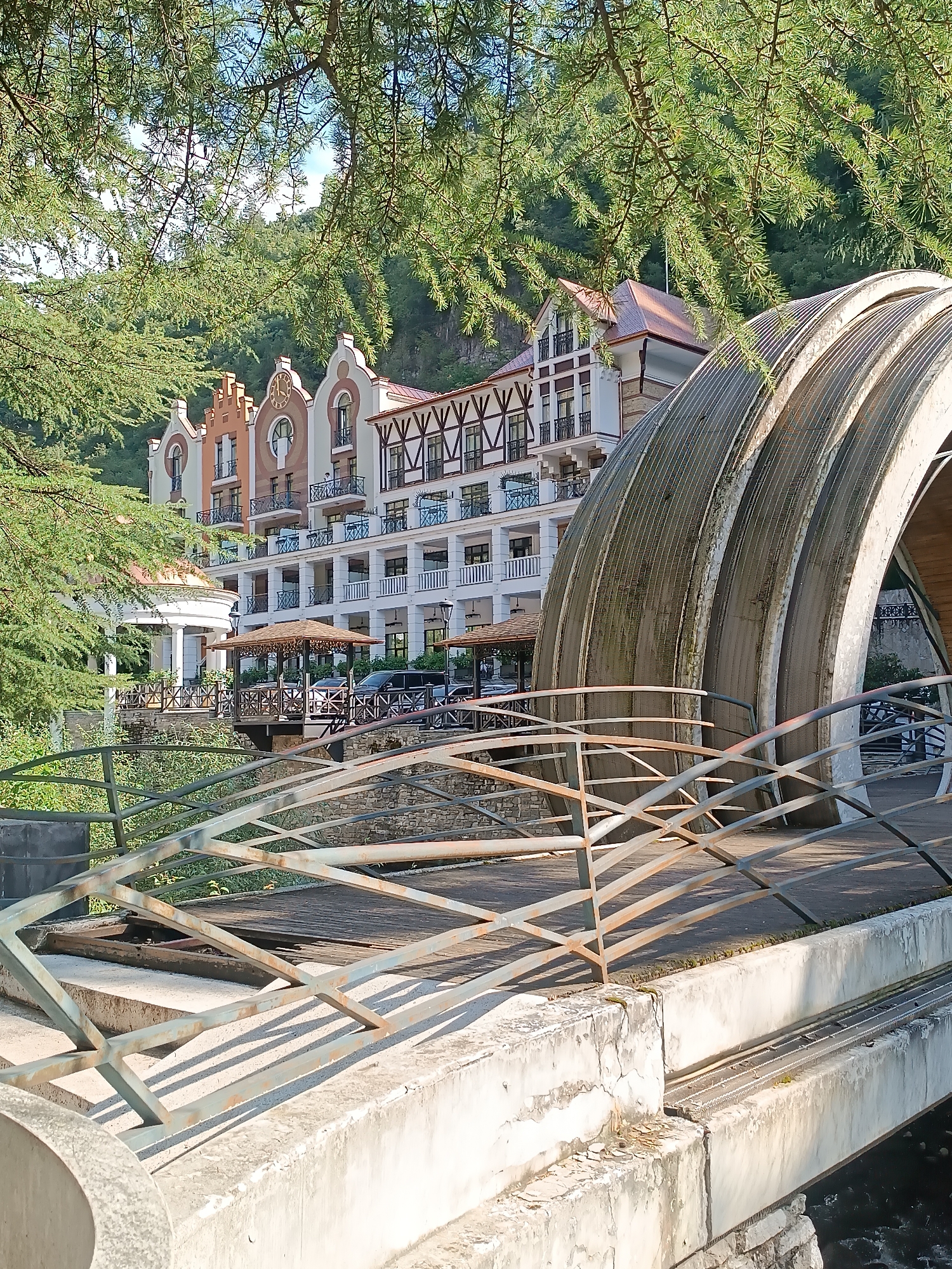 The image shows a view of the Borjomi Central Park in Borjomi, Georgia. In the foreground, a concrete bridge with a metal railing is visible. The railing is aged and shows signs of rust. Beyond the bridge, a large, curved, metallic structure, possibly an awning or part of a building, dominates the right side of the frame. In the background, a multi-story building with ornate architecture, featuring distinctive roof gables, is set against a backdrop of lush green trees and a hillside. The building appears to be a hotel or a similar establishment, with balconies and decorative elements. There are also gazebos and outdoor seating areas visible on the building's terrace. The sky is clear and blue, and sunlight illuminates the scene. The overall impression is one of a resort or park setting with a blend of natural beauty and architectural interest.