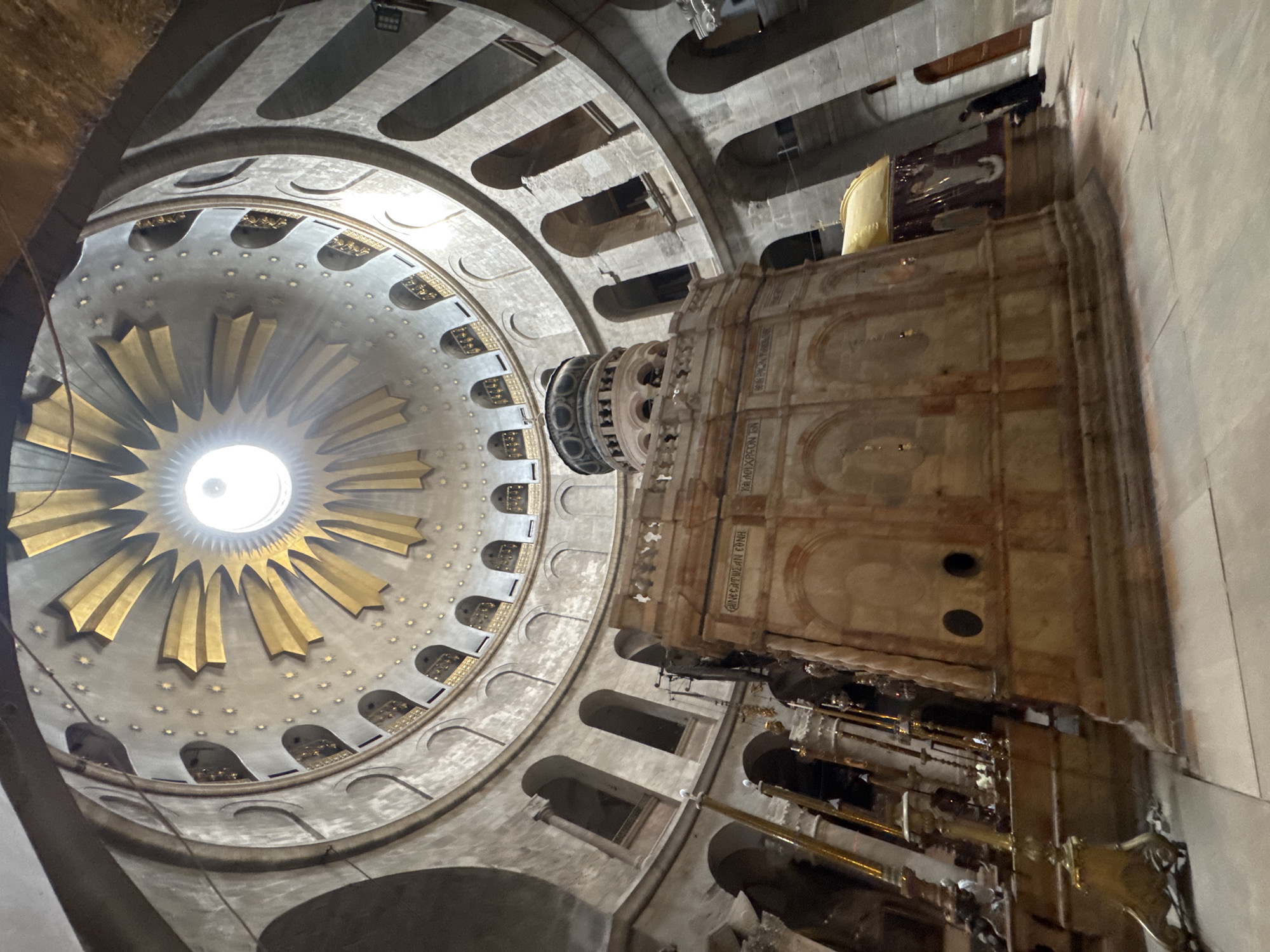 The image depicts the interior of the Church of the Holy Sepulchre in Jerusalem, Israel. The central focus is the Aedicule, a structure believed to enclose the tomb of Jesus. The Aedicule is adorned with pinkish marble and features inscriptions. Above it, the vast rotunda of the church is visible, characterized by its circular design with tiered arches and windows. The ceiling is a domed structure with golden rays emanating from a central oculus, which allows natural light to illuminate the space. Several people are present, including one individual in dark religious attire standing near the Aedicule and others partially visible in the background. The floor is paved with stone. Various religious artifacts, such as candelabras and lamps, are scattered throughout the foreground and around the Aedicule, adding to the sacred atmosphere of the location. The architecture is ancient and imposing, conveying a sense of historical and religious significance.