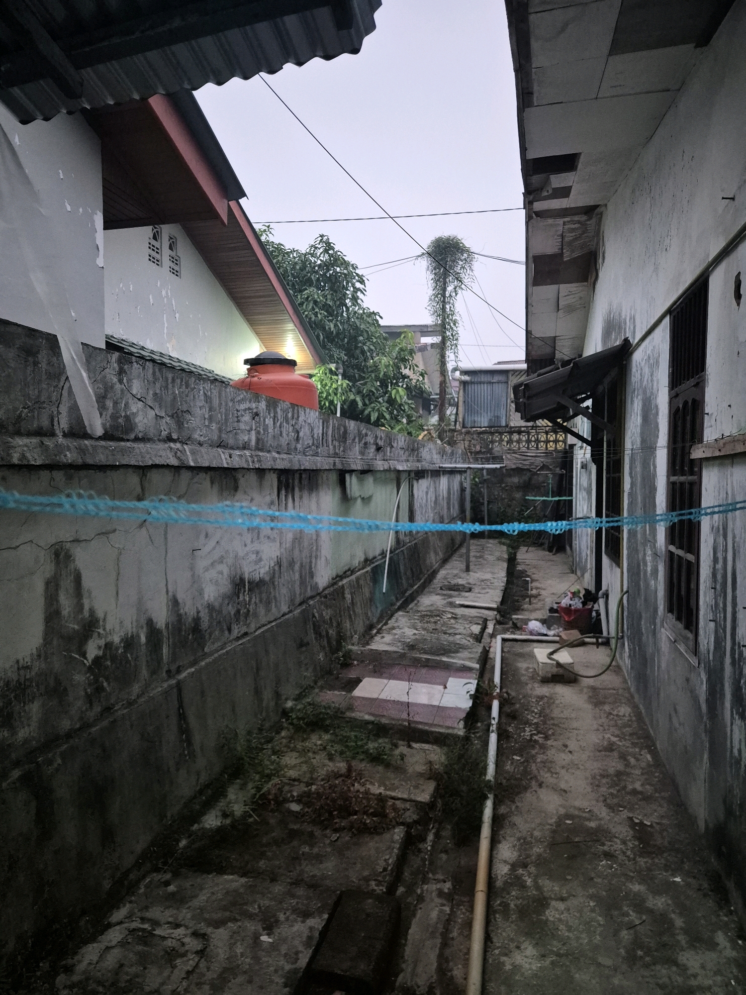 The image depicts a narrow alleyway between two buildings. The left side of the alley is bordered by a high, weathered concrete wall, which appears to be the exterior wall of a residential property. On top of this wall, an orange water tank is visible. The building on the right side of the alley has a rough, white-painted exterior wall with a dark-colored window. A blue rope is strung horizontally across the alleyway. The ground is paved with concrete and there is some vegetation growing in cracks and along the edges. In the background, a tall, slender plant with hanging foliage is visible, along with other structures and trees. The sky is overcast, suggesting it might be early morning or late afternoon. The overall impression is one of a somewhat neglected urban or semi-urban environment. Given the context of Bontang, Indonesia, this scene could represent a back alley in a residential area.