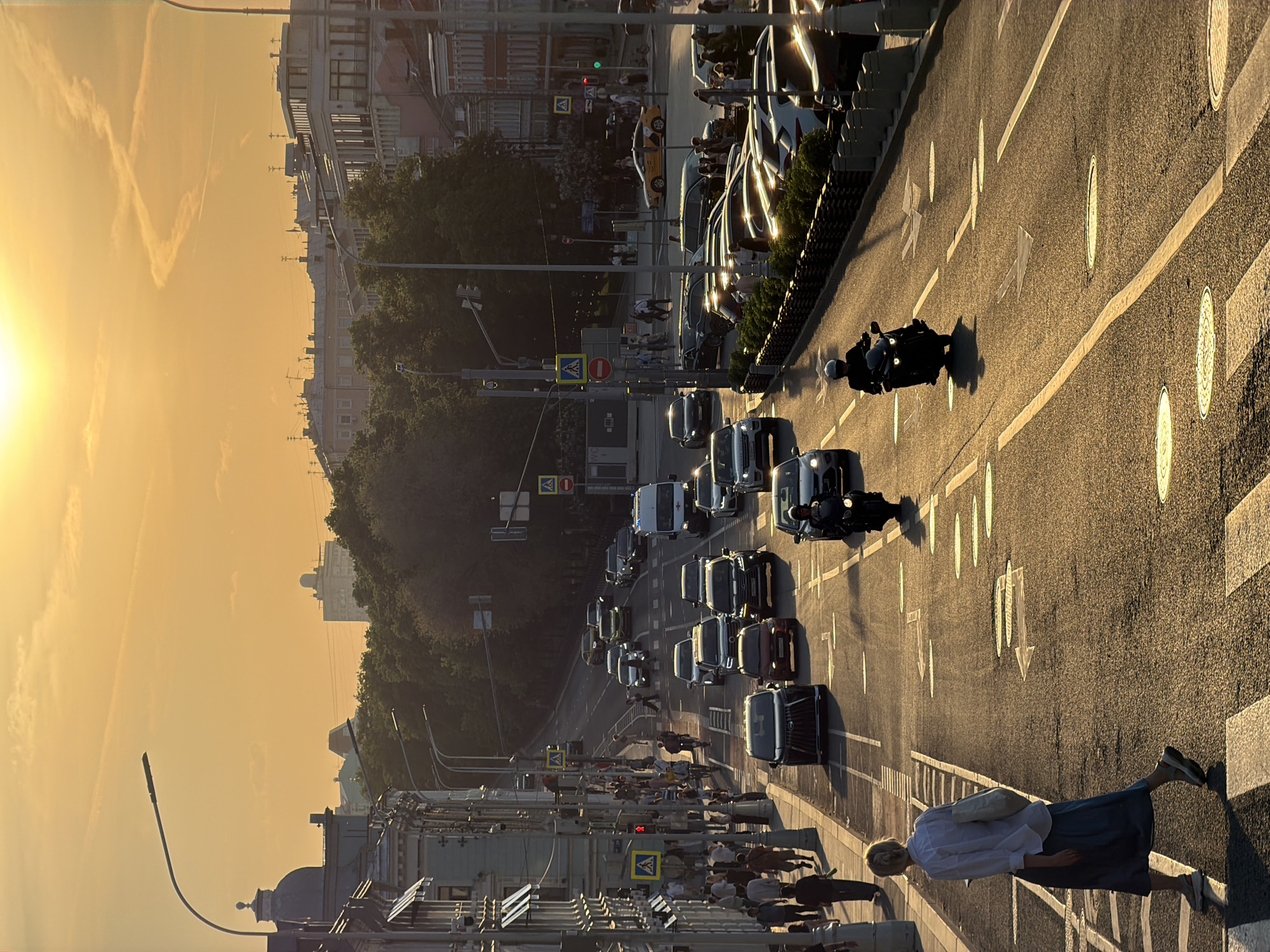 The image depicts a street scene in Moscow, Russia, during sunset. The shot, taken from an elevated vantage point, shows a wide street with multiple lanes of traffic, including cars and motorcycles. Pedestrians are visible on the sidewalks and crosswalks. Streetlights and buildings line the sides of the street. The sky is a gradient of orange and yellow due to the setting sun. The street is marked with lane dividers, directional arrows, and pedestrian crosswalks.