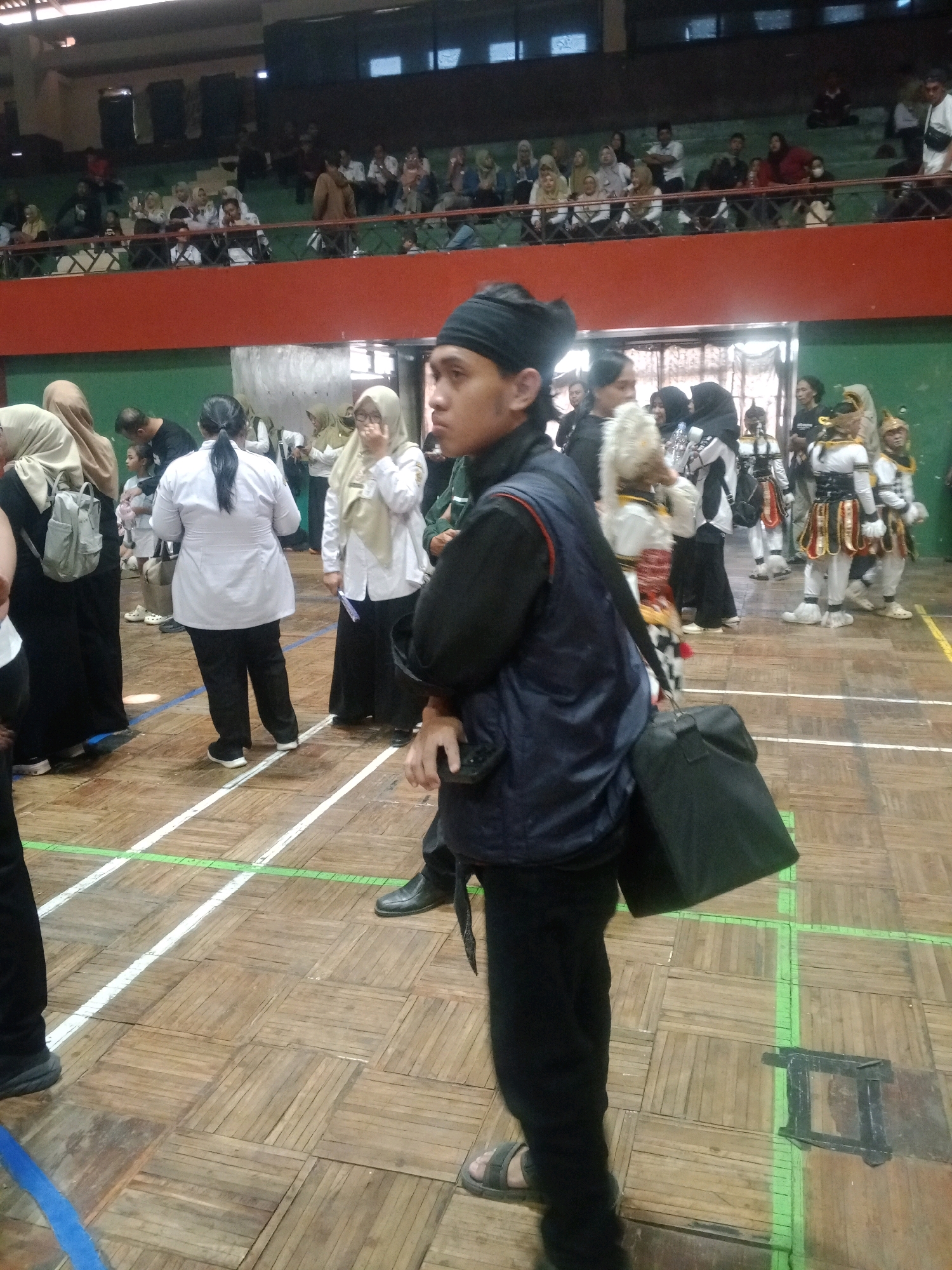 The image depicts an indoor event, likely a competition or performance, taking place in the city of Wuryorejo, Indonesia. The setting appears to be a gymnasium with a polished wooden floor marked with white and green lines, suggesting a sports or performance arena. In the foreground, a young man wearing a black bandana and a dark vest over a black shirt and pants is prominently positioned, facing towards the right of the frame. He is carrying a black bag over his left shoulder.
In the mid-ground and background, various individuals are visible. To the left, a woman in a cream-colored hijab and black clothing stands with a backpack. Further back, other participants and spectators are present. Some individuals are dressed in traditional or ceremonial attire, including those with elaborate headdresses and colorful costumes, suggesting a cultural performance or martial arts demonstration. Several women are wearing hijabs. The background shows tiered seating filled with an audience, indicating a public event. The overall atmosphere seems to be that of an organized gathering or competition.