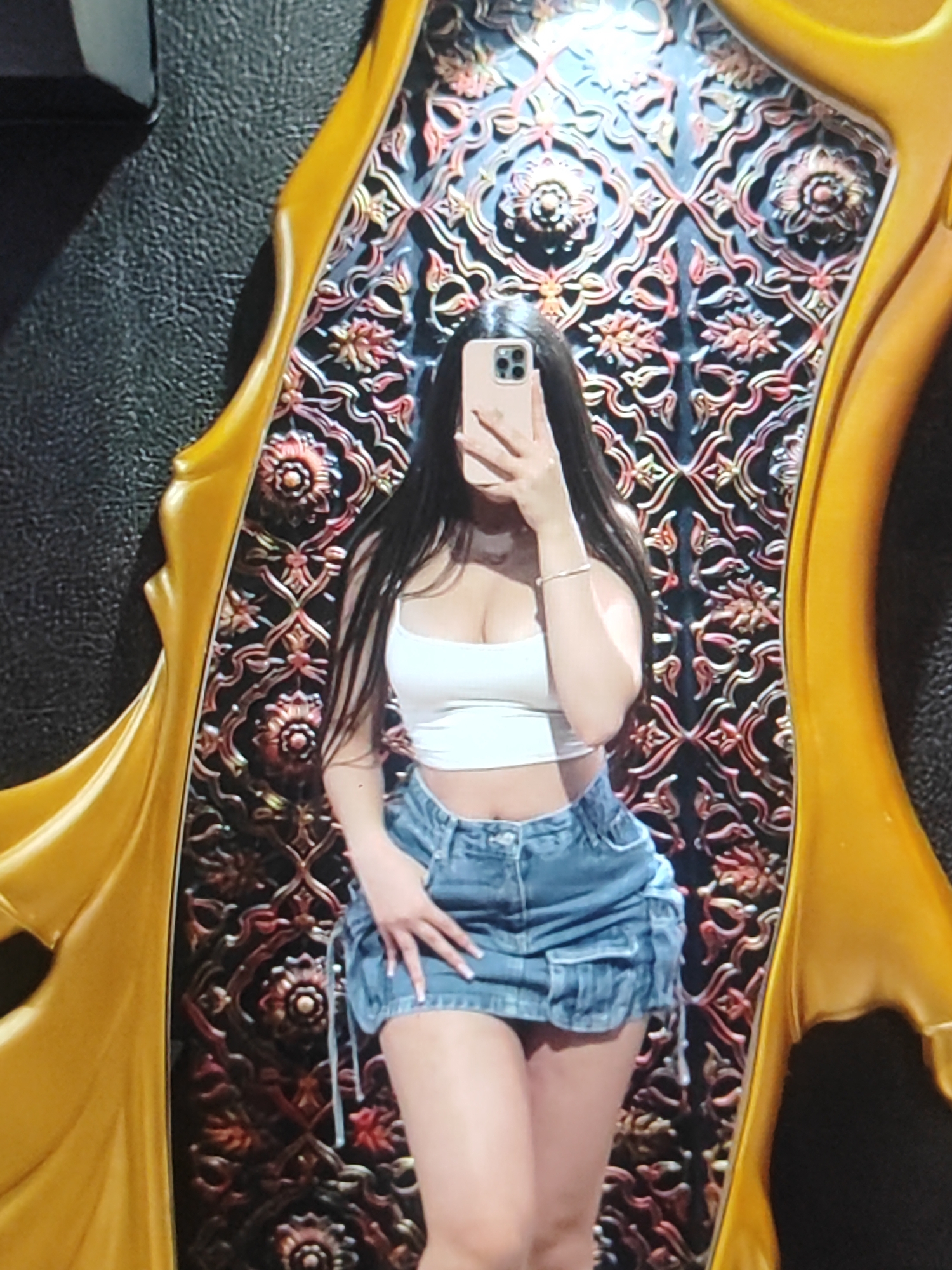 The media file shows a full-body selfie of a young woman taken in what appears to be a room with ornate decor. She is standing in front of a large, gold-framed mirror with a highly decorative, dark background featuring intricate floral and geometric patterns in red and gold hues. The woman is wearing a white crop top and a denim mini skirt. Her hair is long and dark, and she is holding a smartphone up to take the picture, partially obscuring her face. Her right hand is resting on her hip, and her left hand is visible near her phone. The lighting suggests an indoor setting, possibly at night or in a dimly lit area, given the reflections and shadows. There is no visible text in the image. The setting, with its elaborate mirror and textured wall, hints at a potentially luxurious or culturally rich environment, fitting with the provided location context of Fergana City, Uzbekistan. The overall mood seems casual and posed for a social media post.